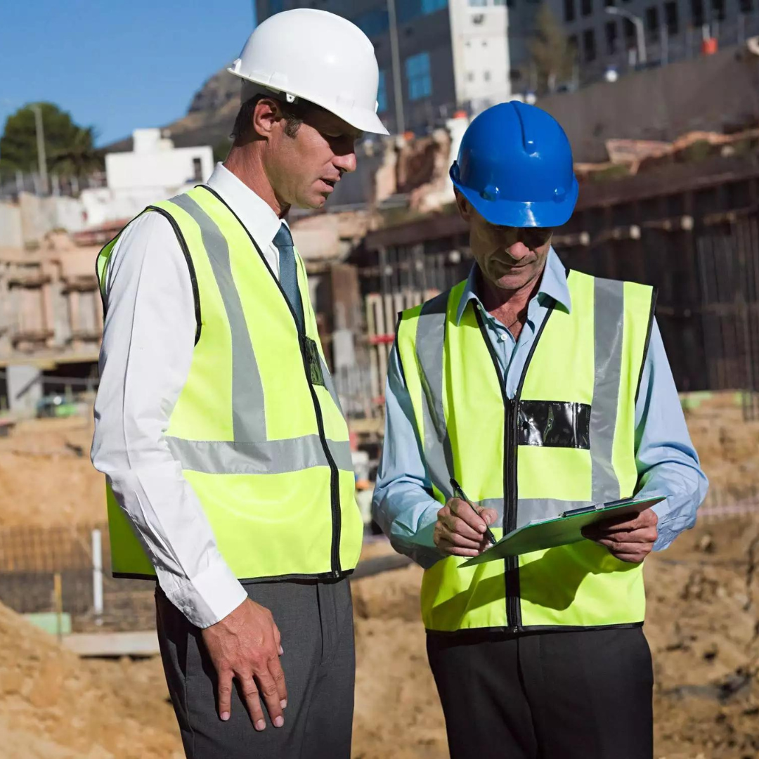 Two men wearing hard hats and safety vests are looking at a clipboard
