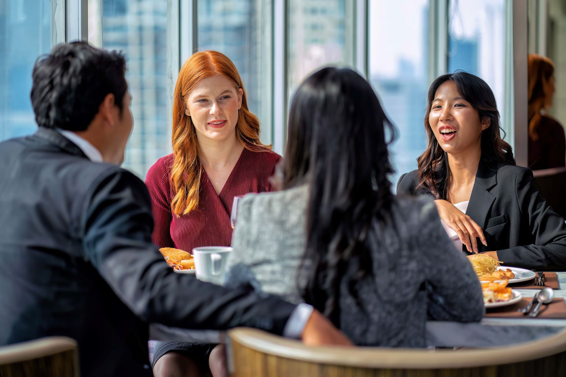Four people at a table, two looking at each other, laughing. City backdrop visible.