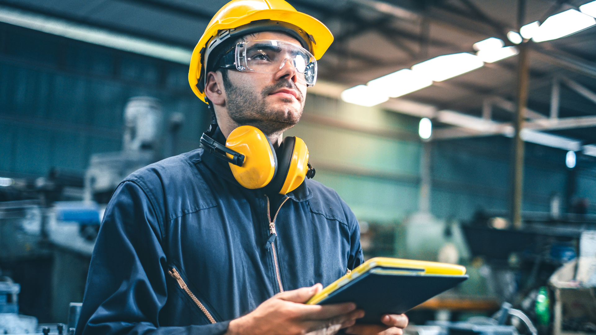A man wearing a hard hat and headphones is holding a tablet in a factory.