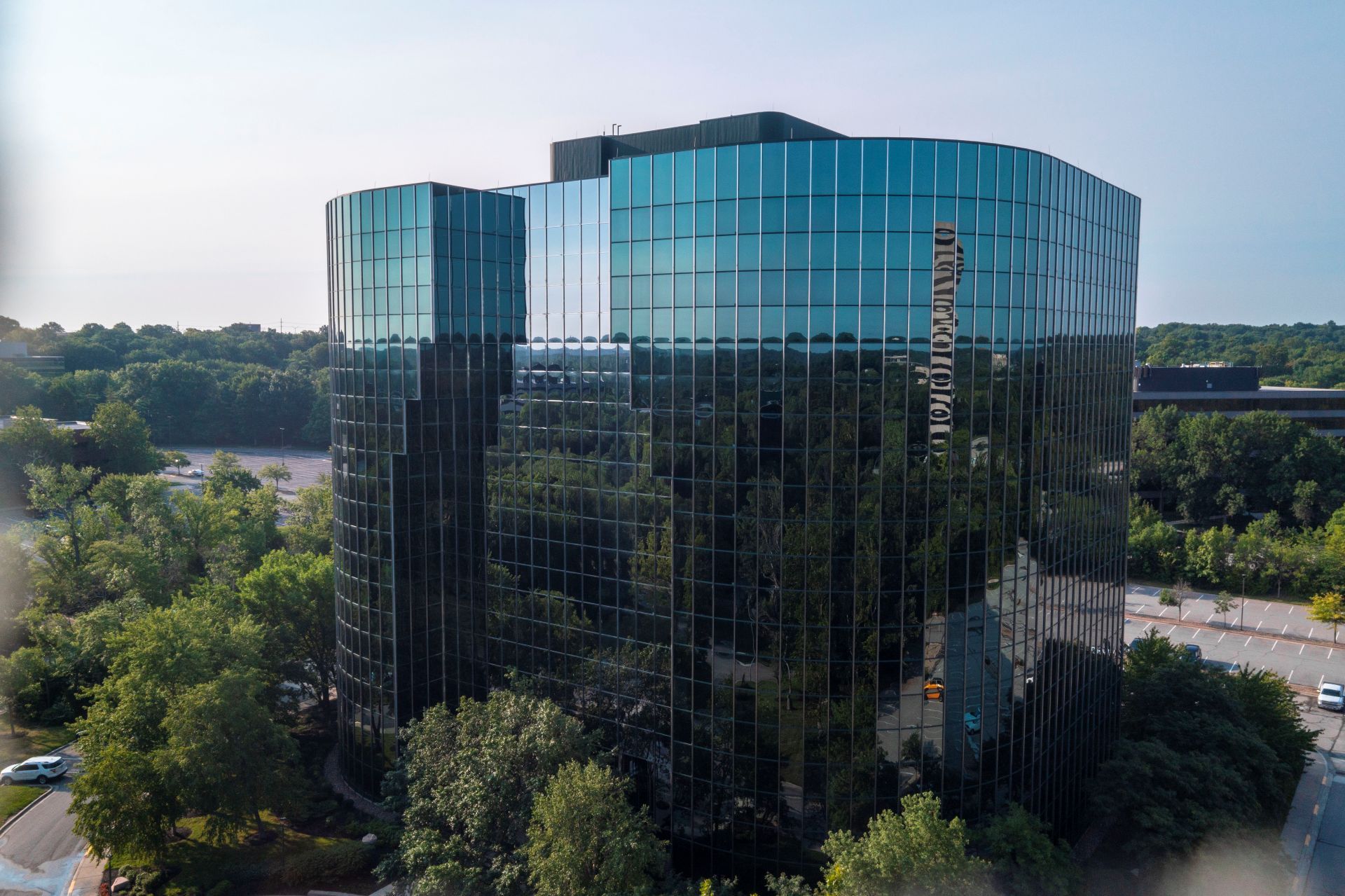 Glass office building surrounded by trees, reflecting a bright sky and city skyline.