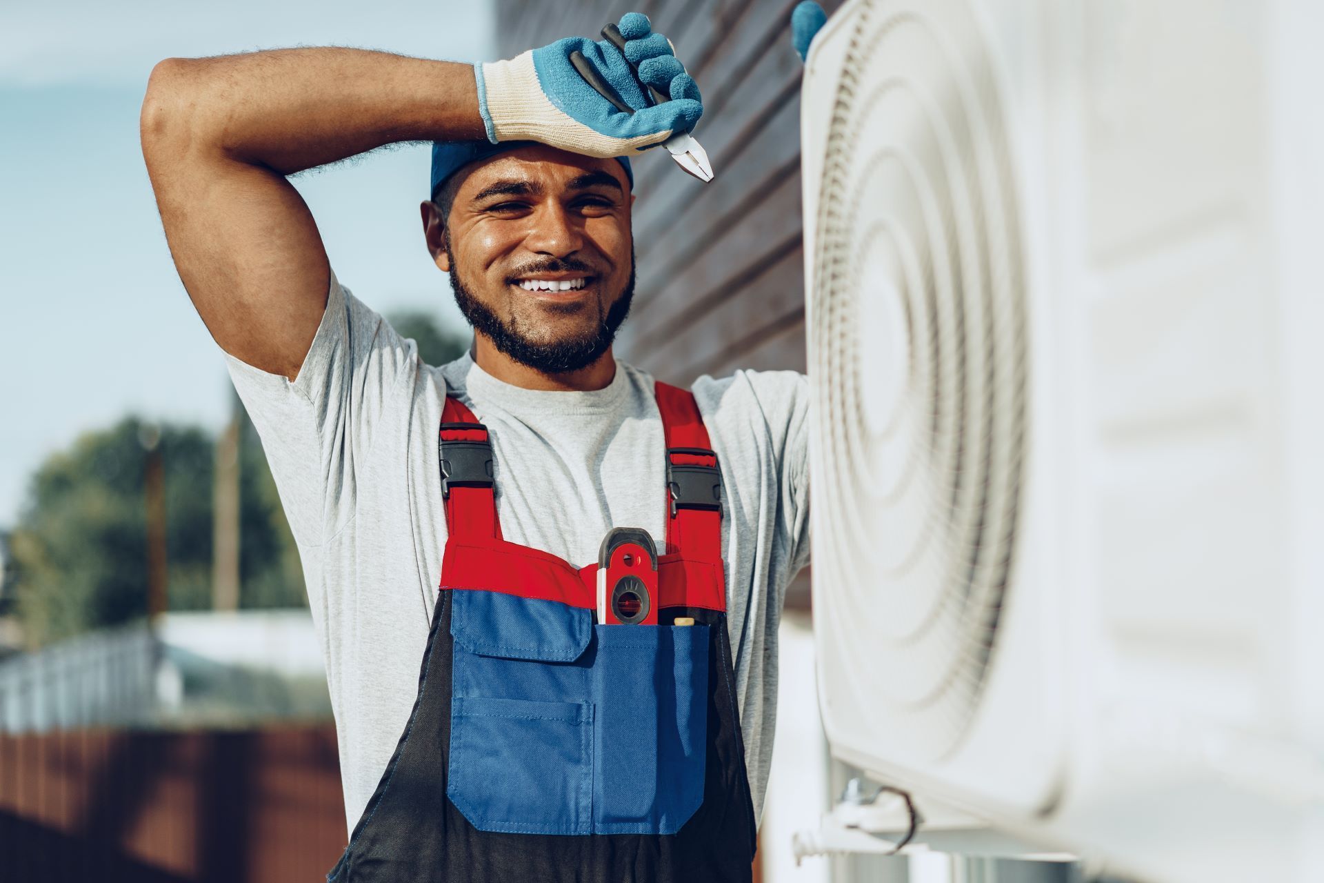Smiling HVAC technician in overalls, outdoors, working on an AC unit.