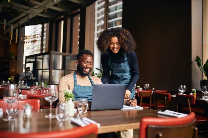 A man and a woman are sitting at a table in a restaurant looking at a laptop.