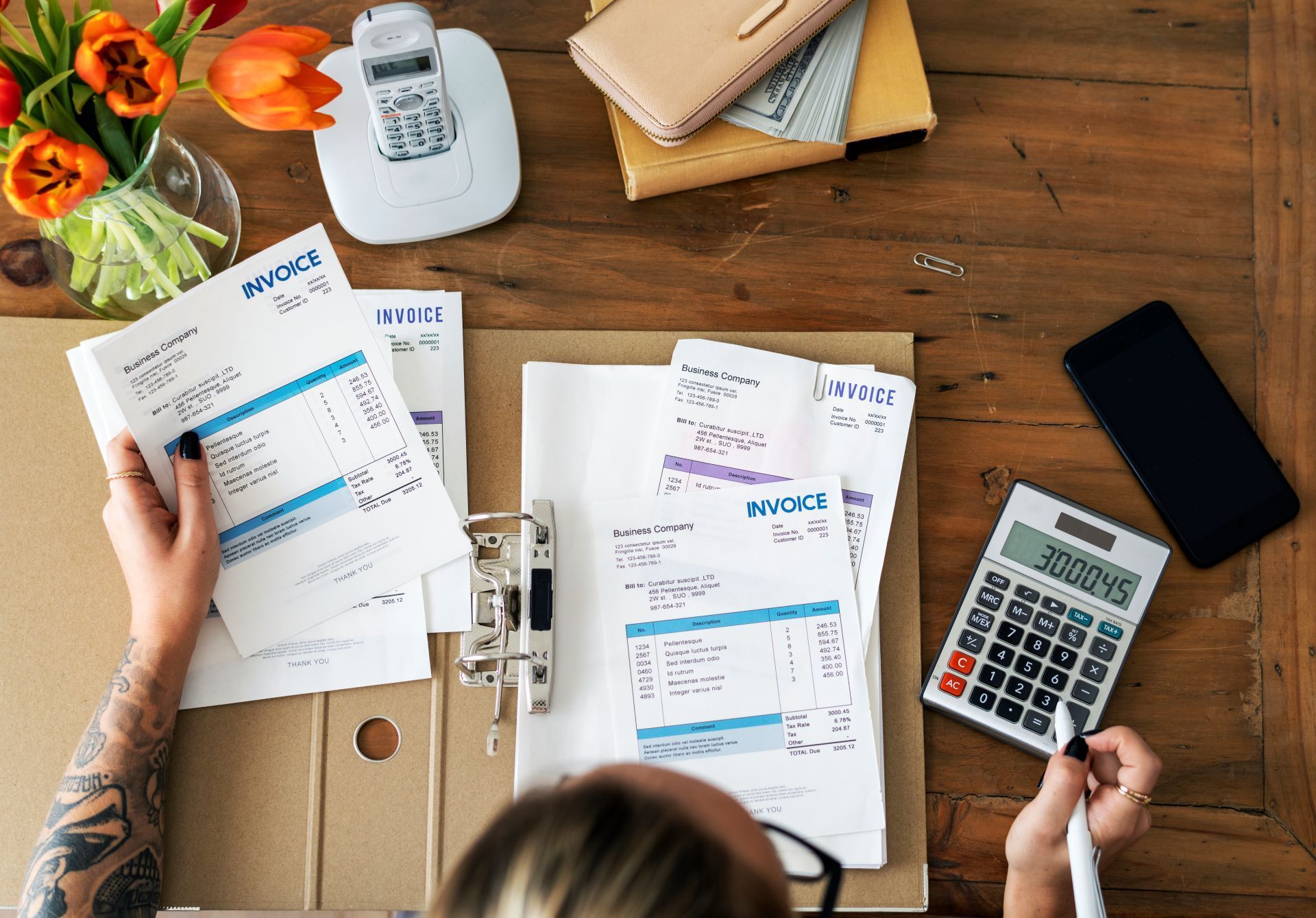 Person reviewing invoices at a desk with calculator, phone, flowers, wallet, and telephone.