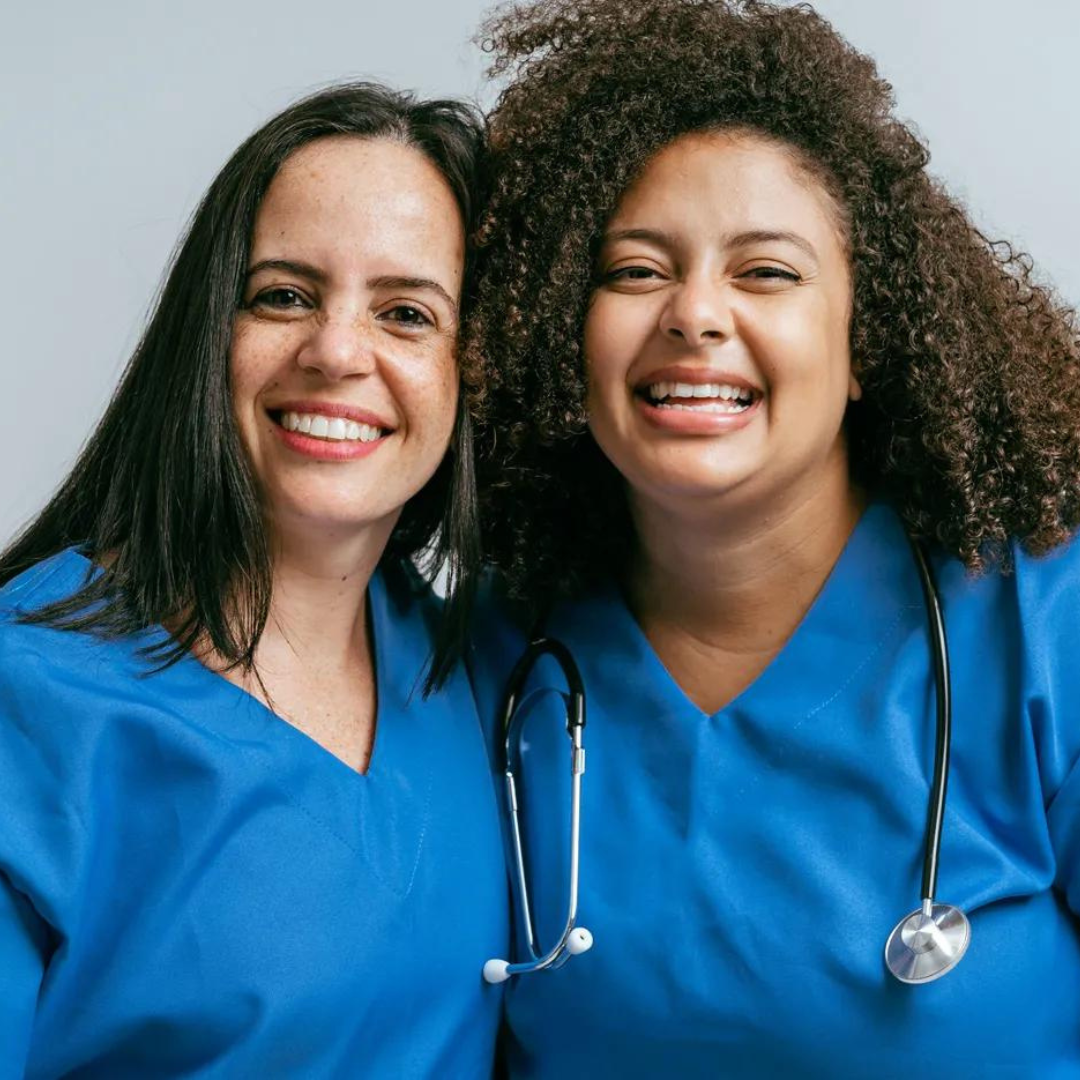 Two nurses wearing blue scrubs and stethoscopes are posing for a picture.