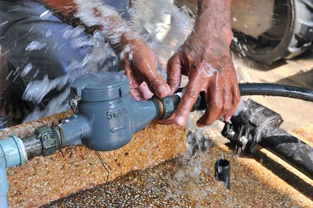 Hands adjusting a blue water pipe valve with water spraying in a work area