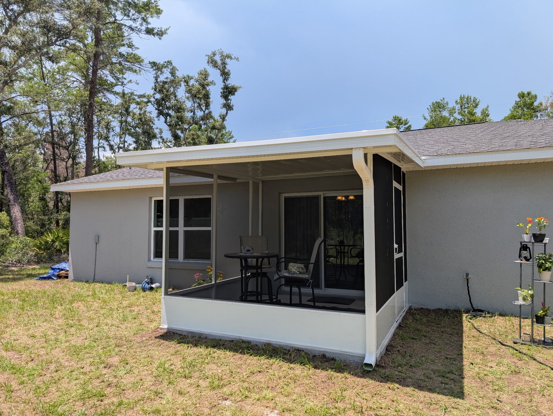 Screened-in patio attached to a light gray house, with seating and a view of trees under a bright blue sky.