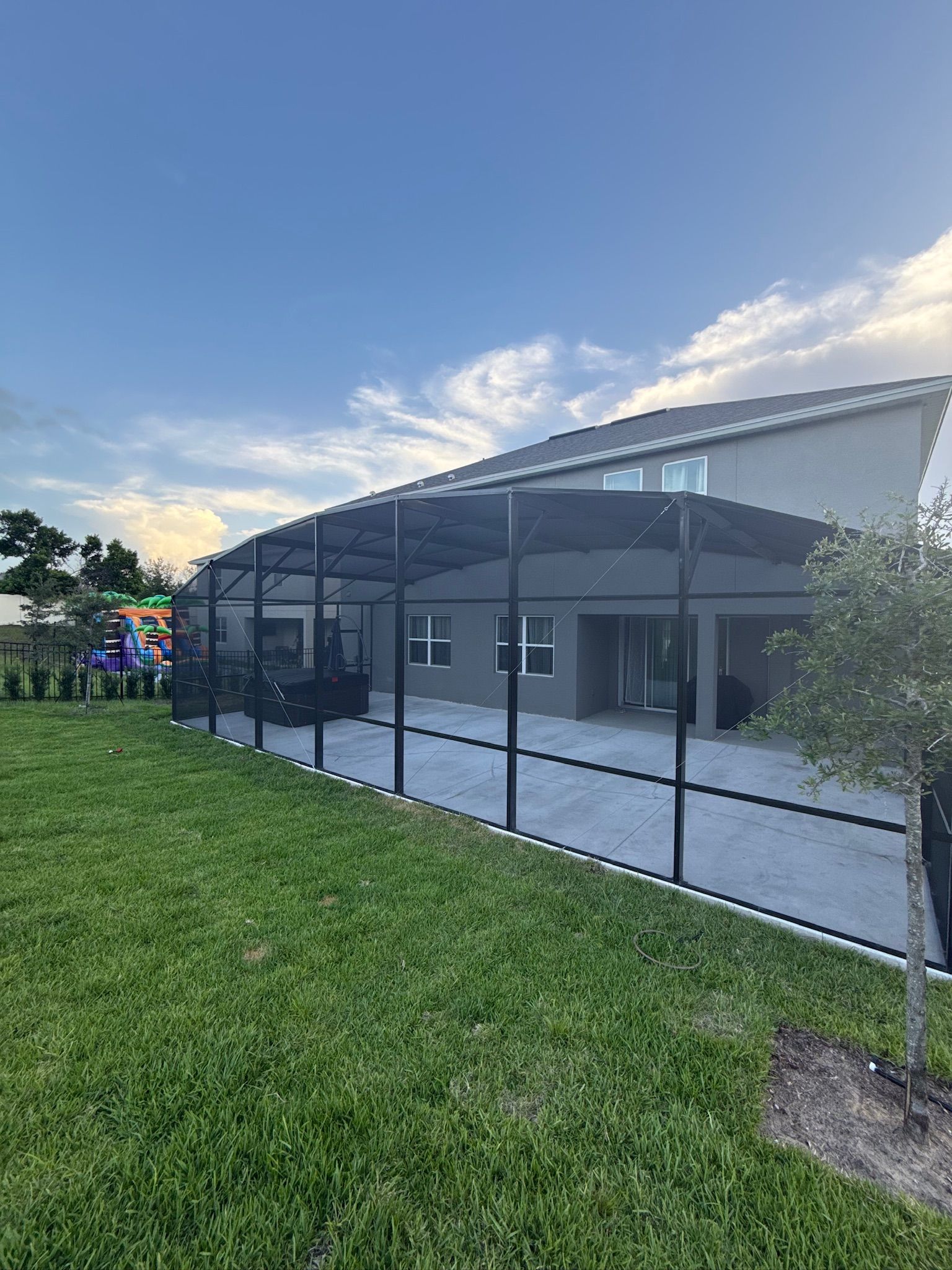 Black fence encloses a backyard with a covered patio, grass, and a two-story house under a blue sky.