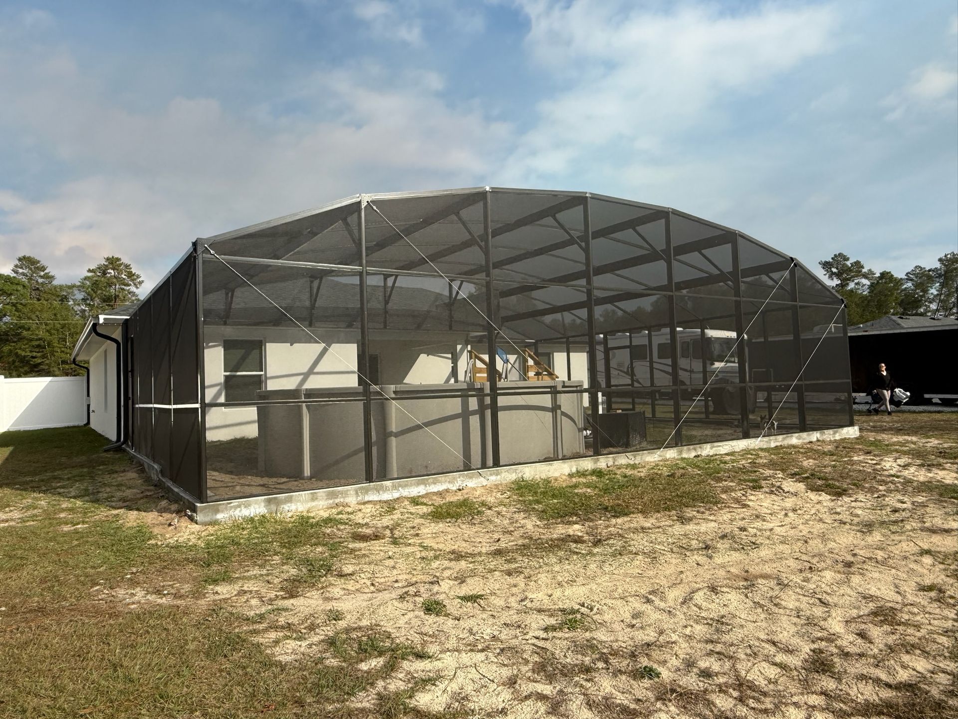 Pool enclosure with arched roof, gray mesh, and a pool on a sandy lot.