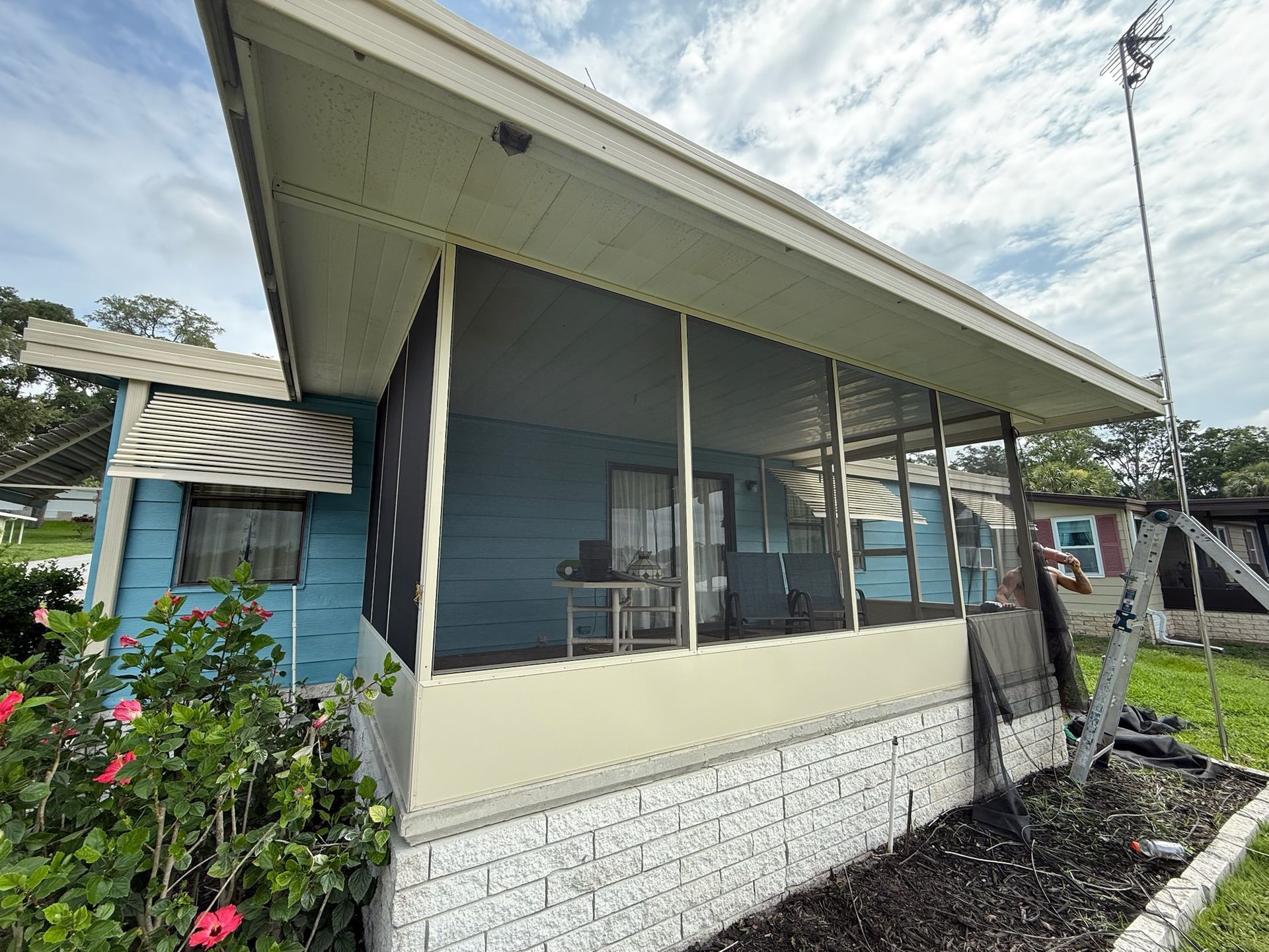 Blue house with screened porch, beige trim. Awnings and landscaping visible. Cloudy sky overhead.