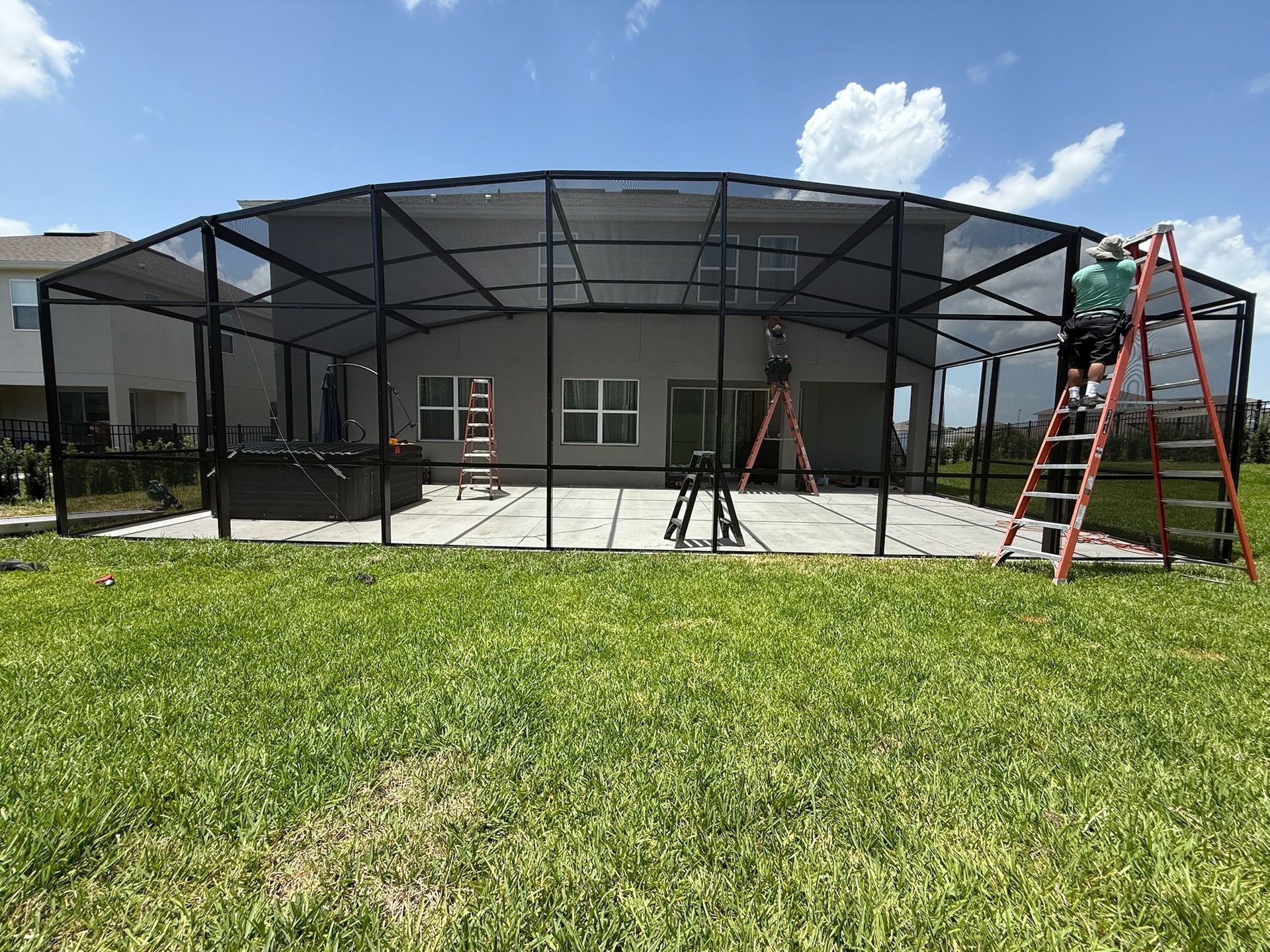 A worker on a ladder installs a black screen enclosure around a patio in a backyard, blue sky.