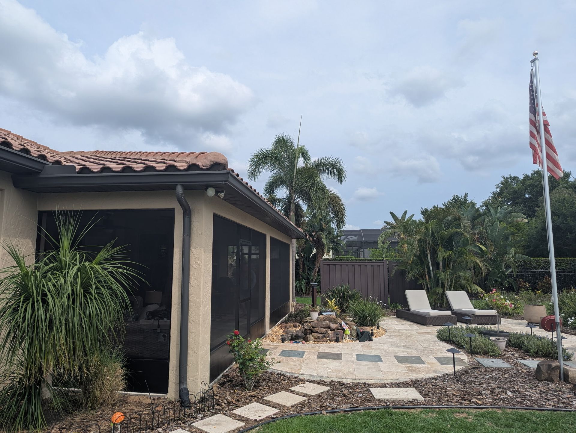 Backyard with a screened patio, flag, stone pathway, and lounge chairs under a cloudy sky.