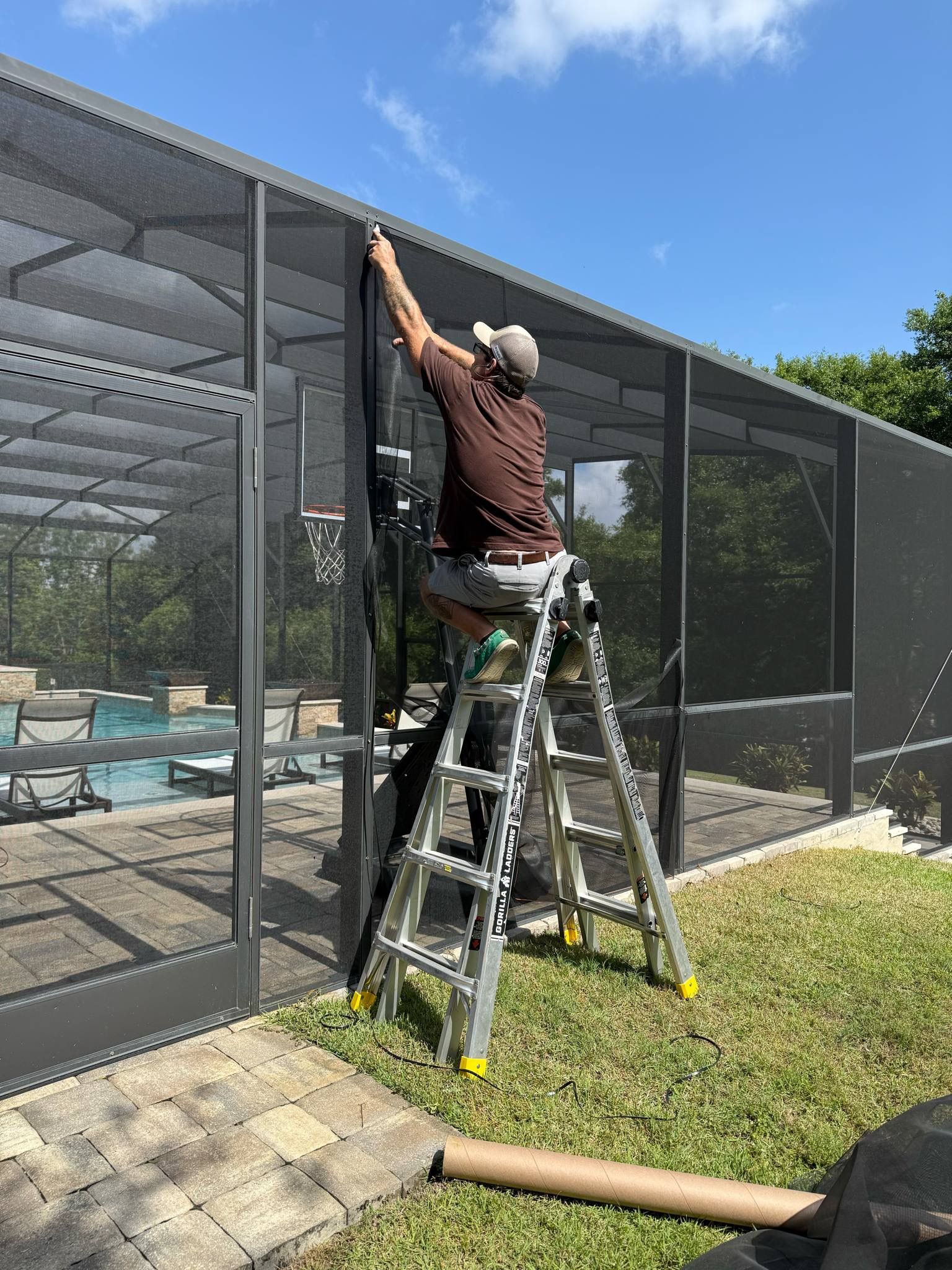 A man is standing on a ladder working on a screened in pool area.