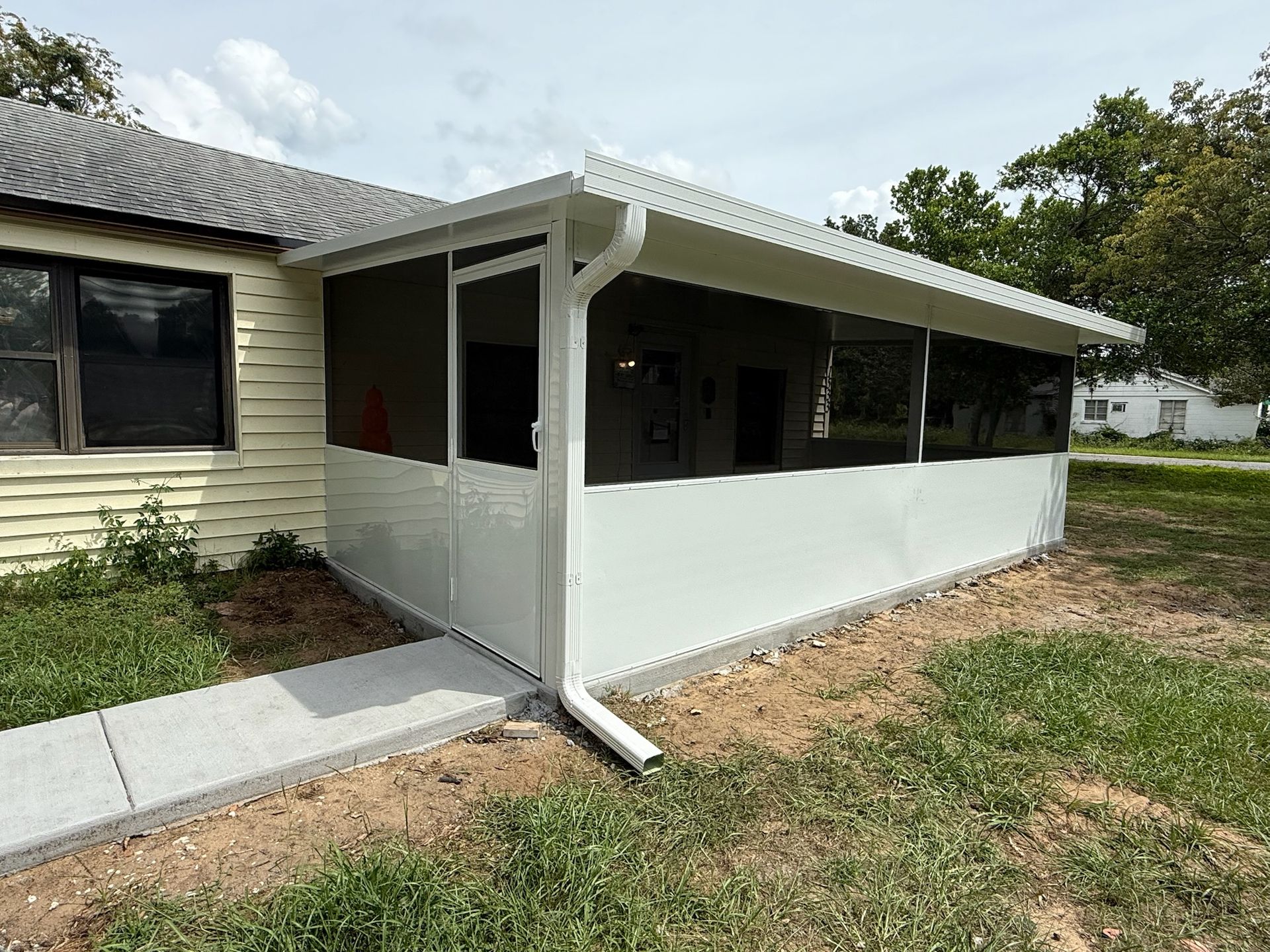 White screened-in porch addition attached to a yellow house with a concrete walkway in a yard.