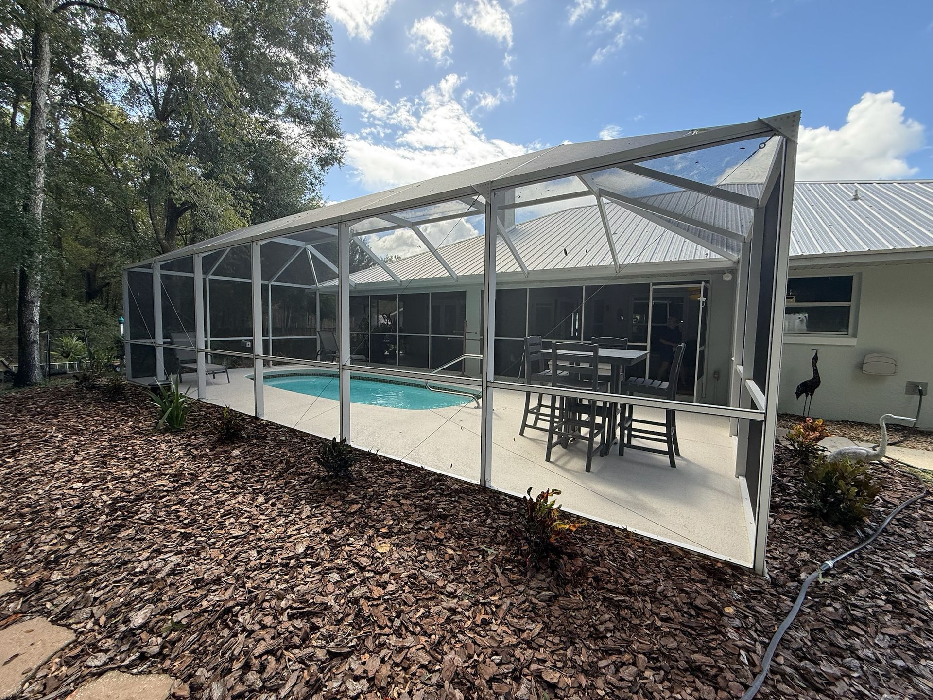 Screened-in pool enclosure with pool, patio furniture, and house in background; sunny day.