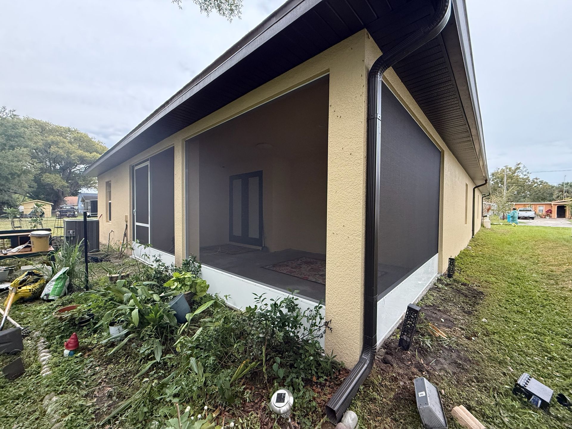Tan stucco home with screened-in porch and black gutters, surrounded by overgrown plants and grass.