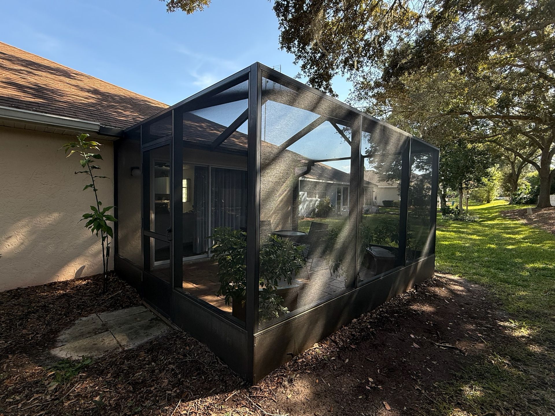 Screened enclosure attached to a house with plants inside and a grassy backyard.