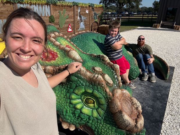 Woman smiles, touches a green dragon sculpture. Two people pose behind it. Outdoor setting, sunny.
