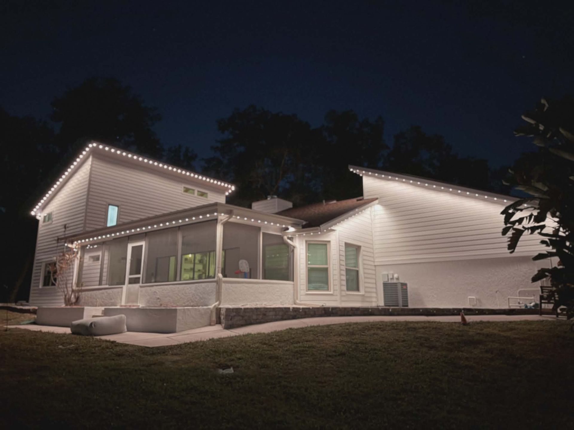 White house exterior at night, lit with string lights. Porch, lawn, and dark sky visible.