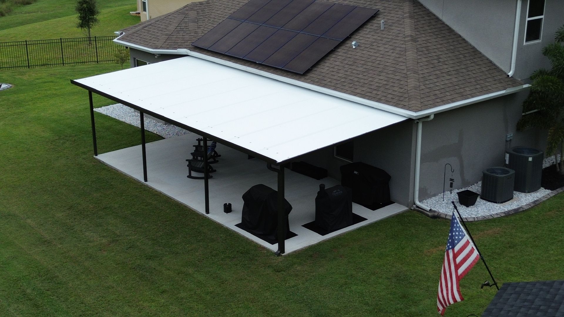 Backyard patio with white roof, black supports, and grill covered in black. American flag in yard.