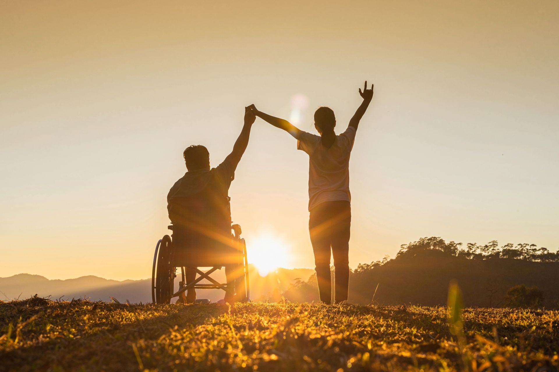 Person in wheelchair and person standing, holding hands, arms raised against a sunset backdrop.