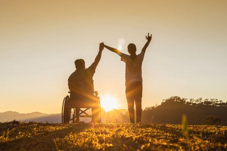 Person in wheelchair and person standing, holding hands, arms raised against a sunset backdrop.