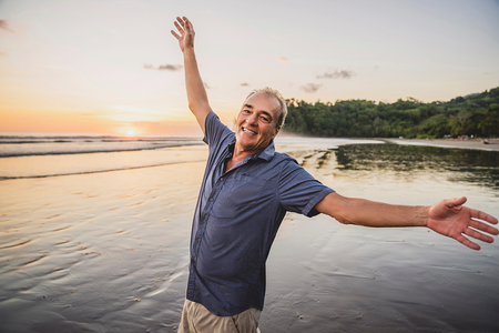 An older man is standing on the beach with his arms outstretched.