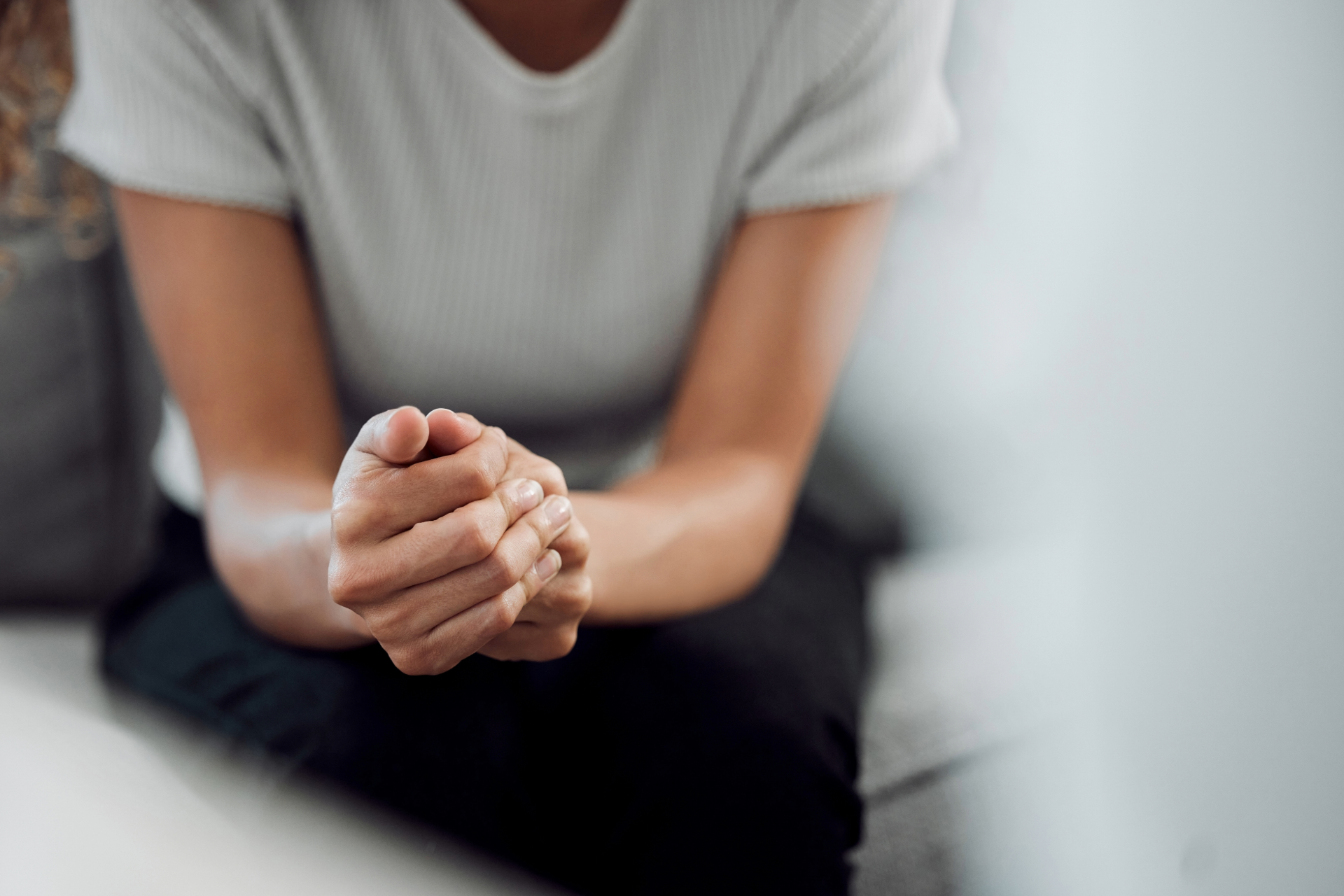 Person sitting with clasped hands in a white shirt and dark pants on a couch