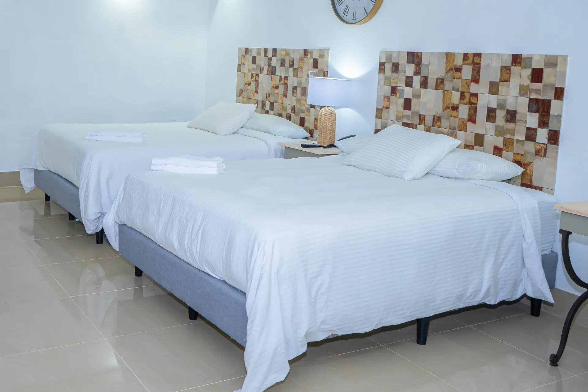 Dos camas en una habitación luminosa con cabeceros de madera, sábanas blancas y reloj.