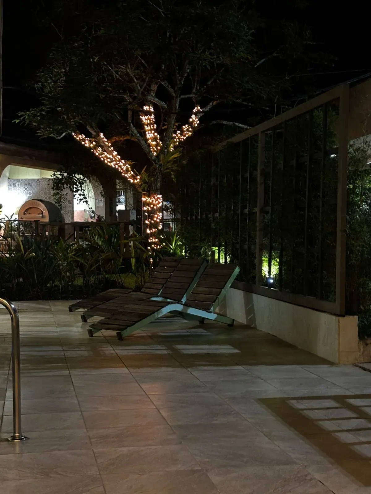 Dos tumbonas se encuentran cerca de un árbol adornado con luces; al fondo hay un muro bajo y un edificio.