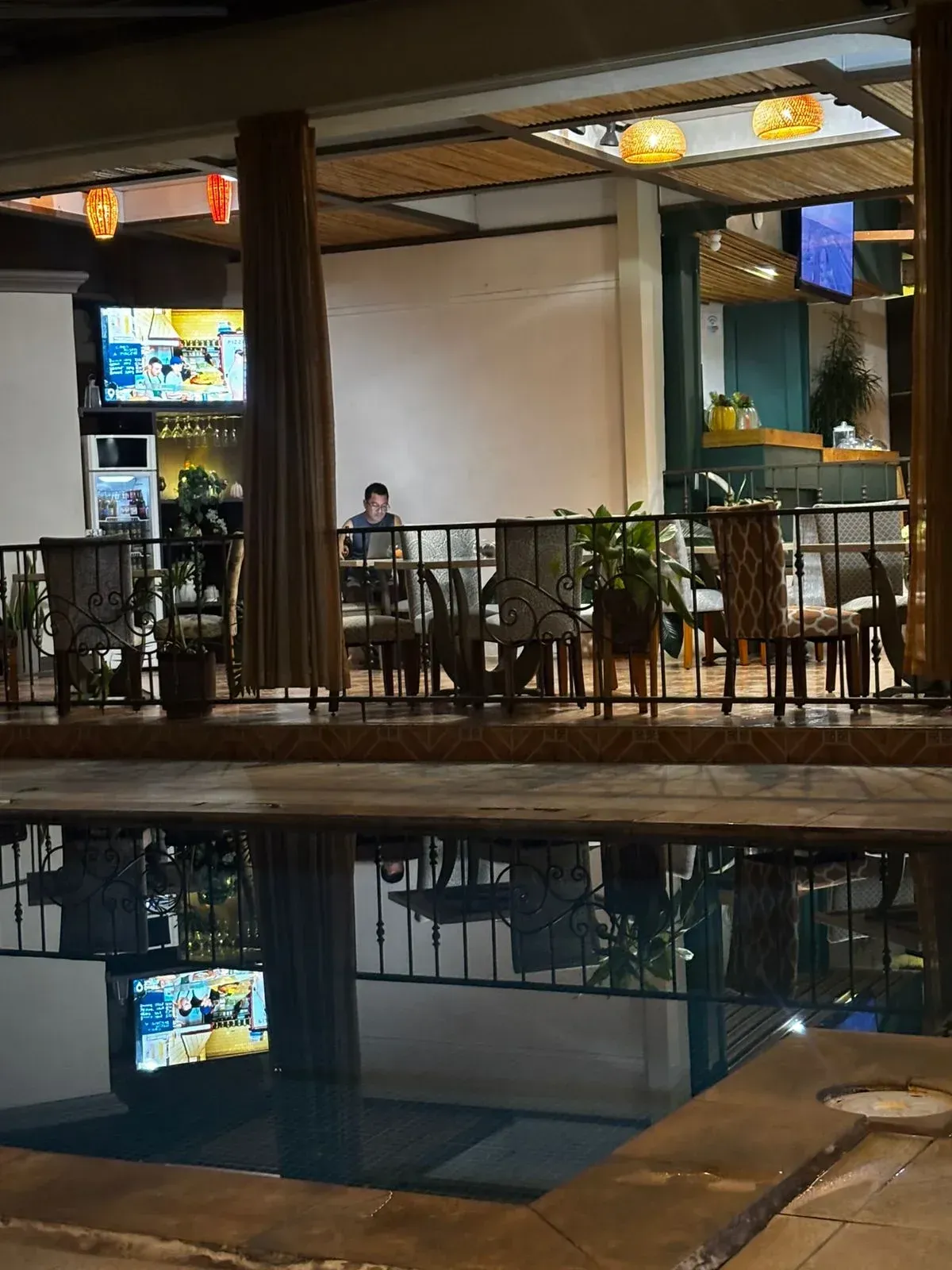 Restaurante junto a la piscina por la noche con agua reflectante, mesas y una persona sentada.