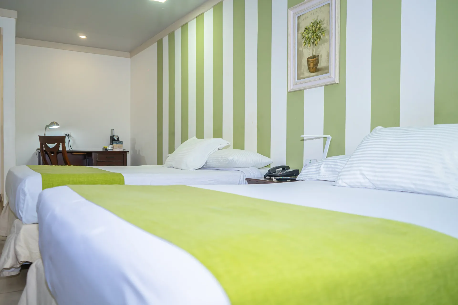 Dos camas en una habitación, con pared de rayas verdes y blancas, escritorio al fondo, pequeño cuadro enmarcado.