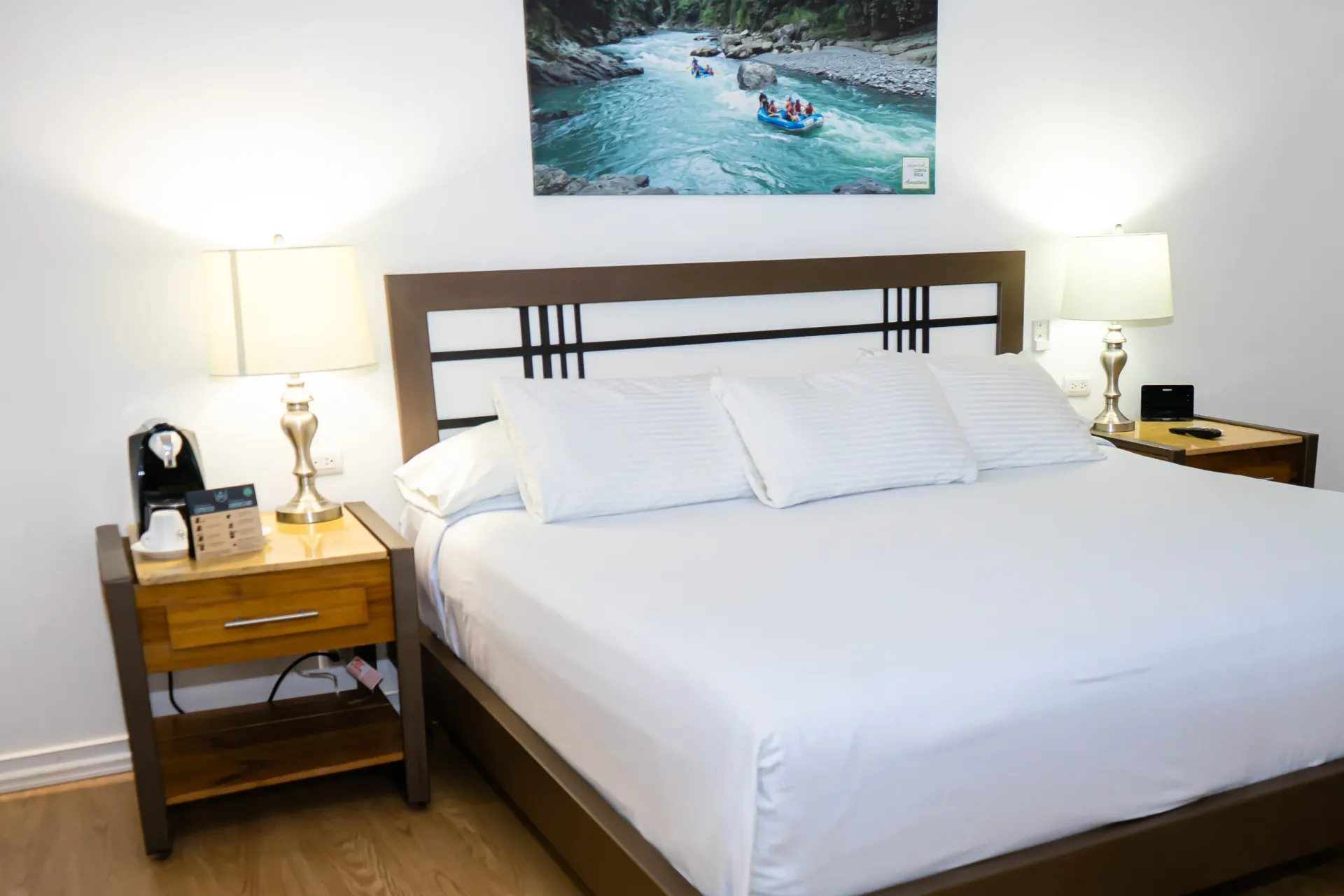 Dormitorio con cama tamaño king, dos lámparas en mesitas de noche, obras de arte sobre la cabecera y cafetera.