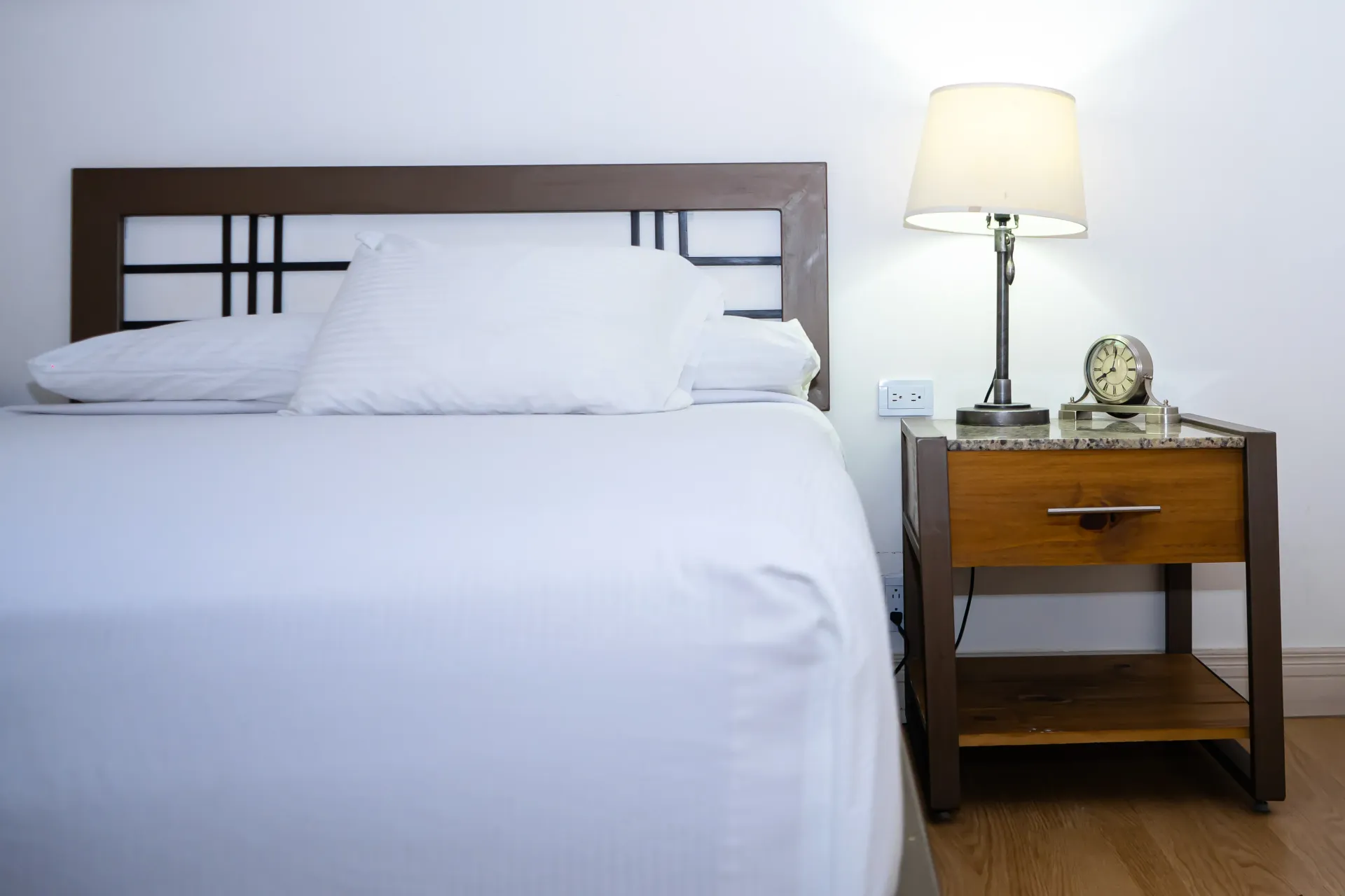 Cama con sábanas blancas y cabecero de marco oscuro, al lado de una mesita de noche de madera con lámpara.