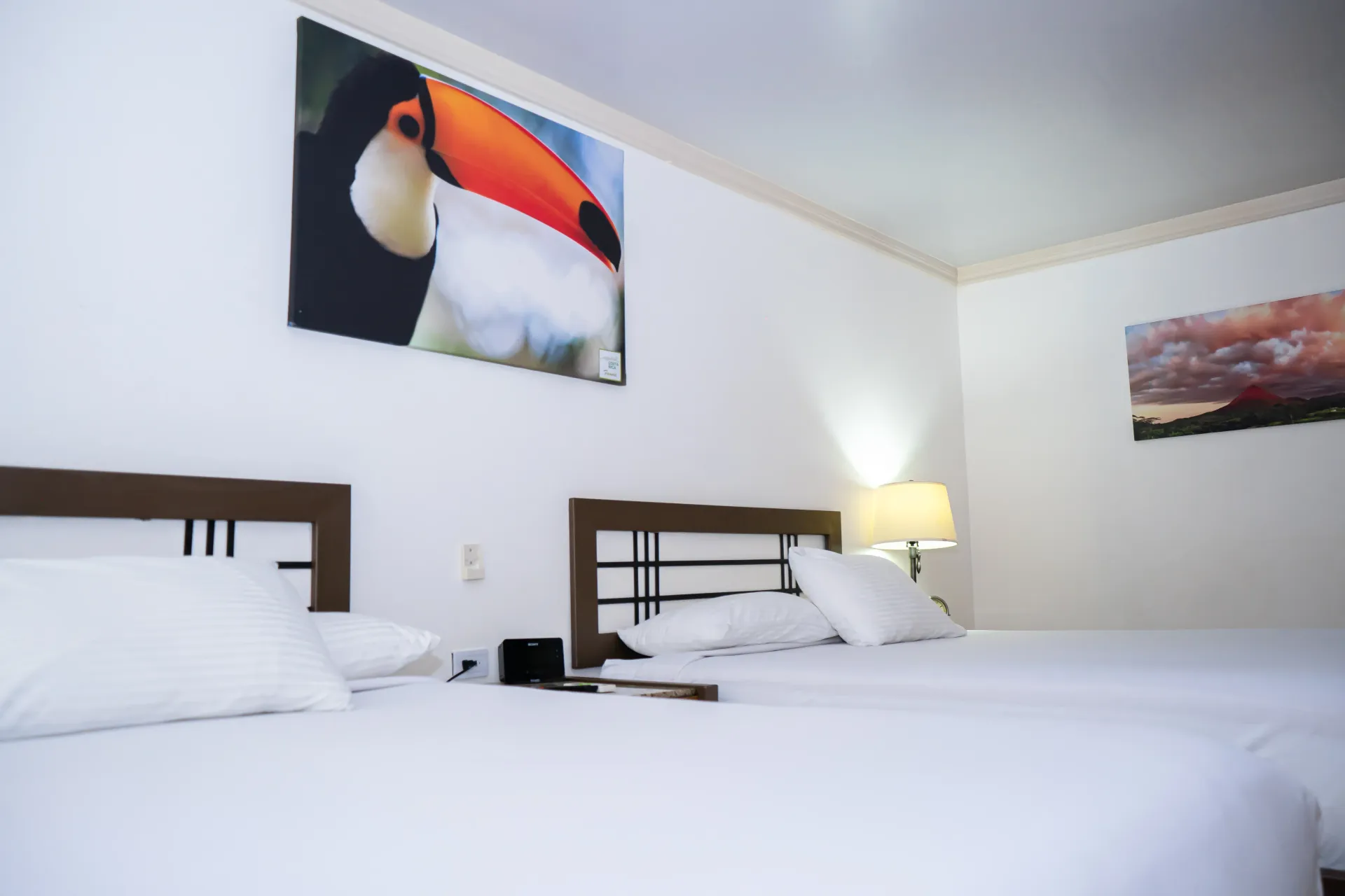 Dos camas en una habitación de paredes blancas con un arte de un tucán y una pintura de un atardecer.