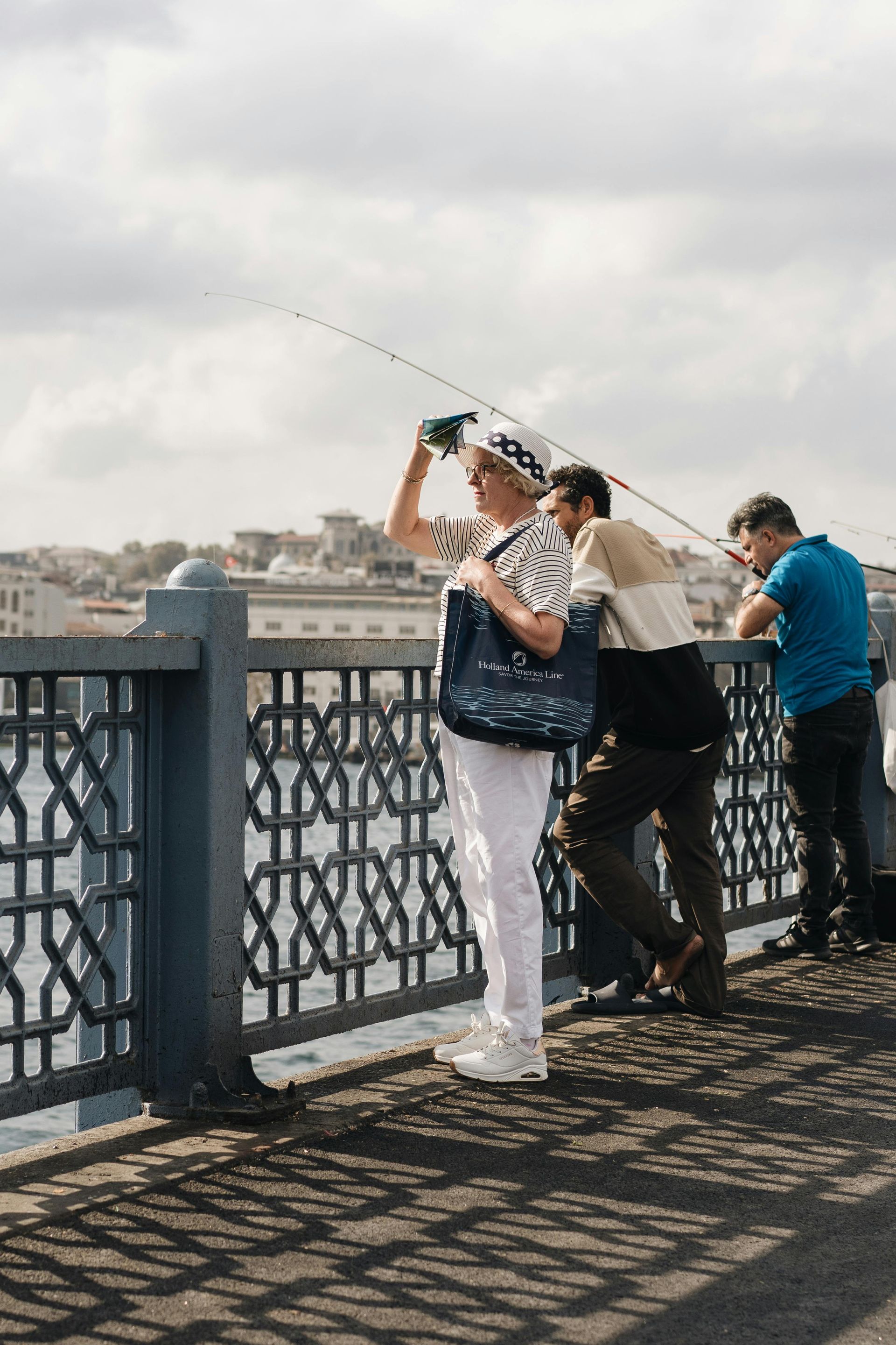 Gente pescando desde un puente. Una mujer con pantalones blancos y sombrero mira hacia el agua. Día soleado.