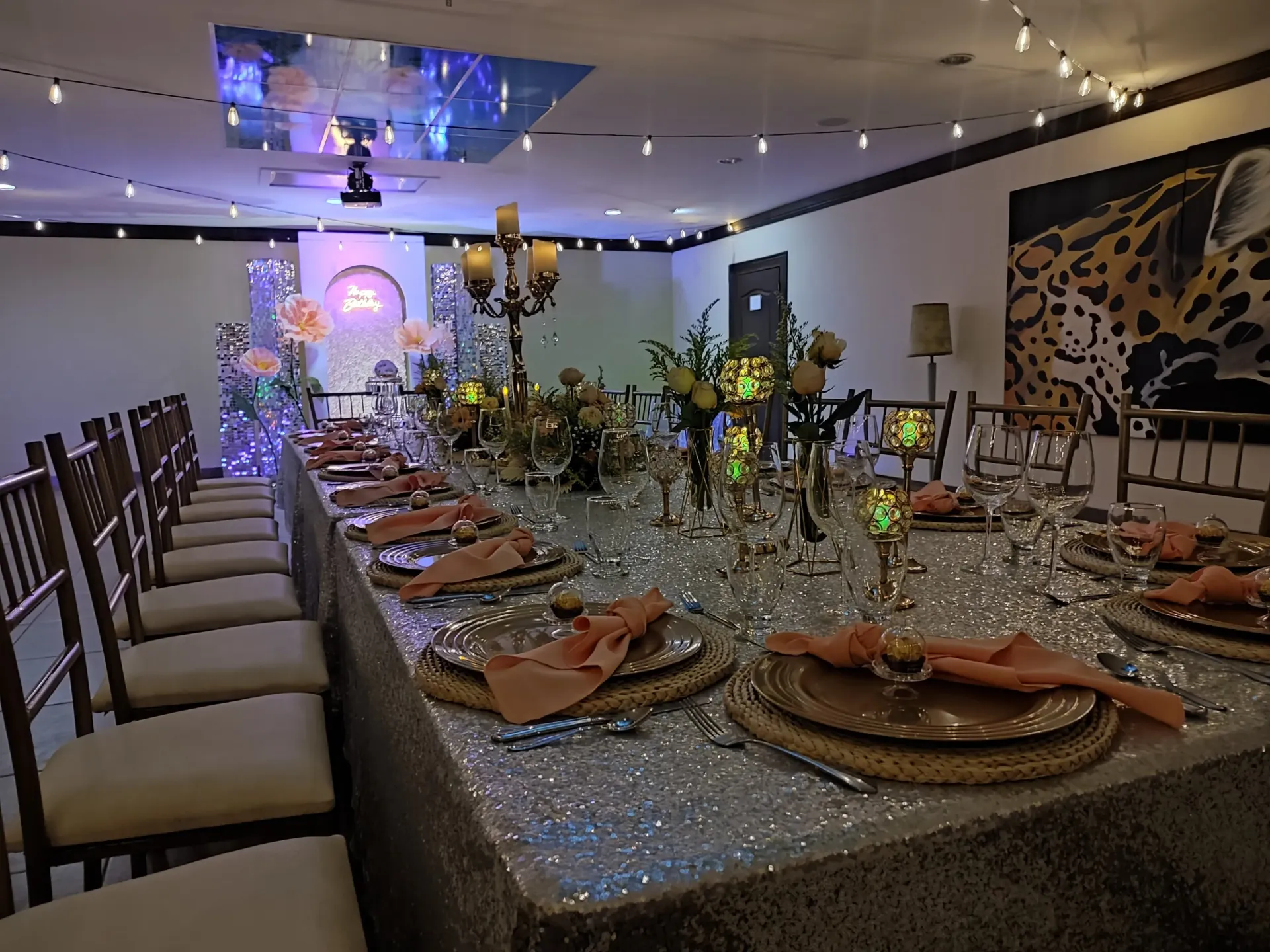 Mesa larga dispuesta para un evento formal con centros de mesa florales y cubiertos ornamentados; luces de cadena y arte