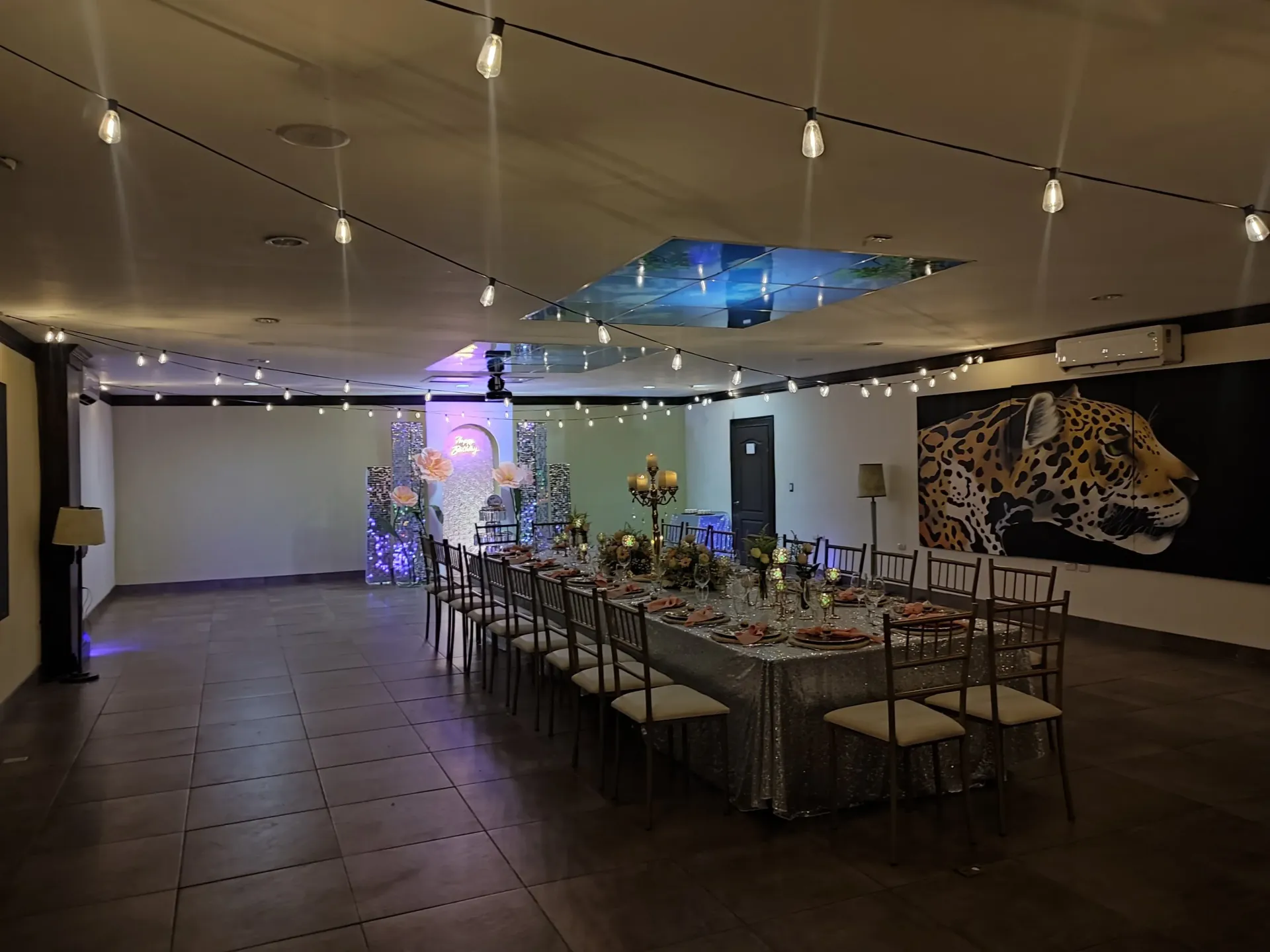 Elegante comedor, mesa larga preparada para una celebración, luces de cadena en el techo, arte de leopardo.