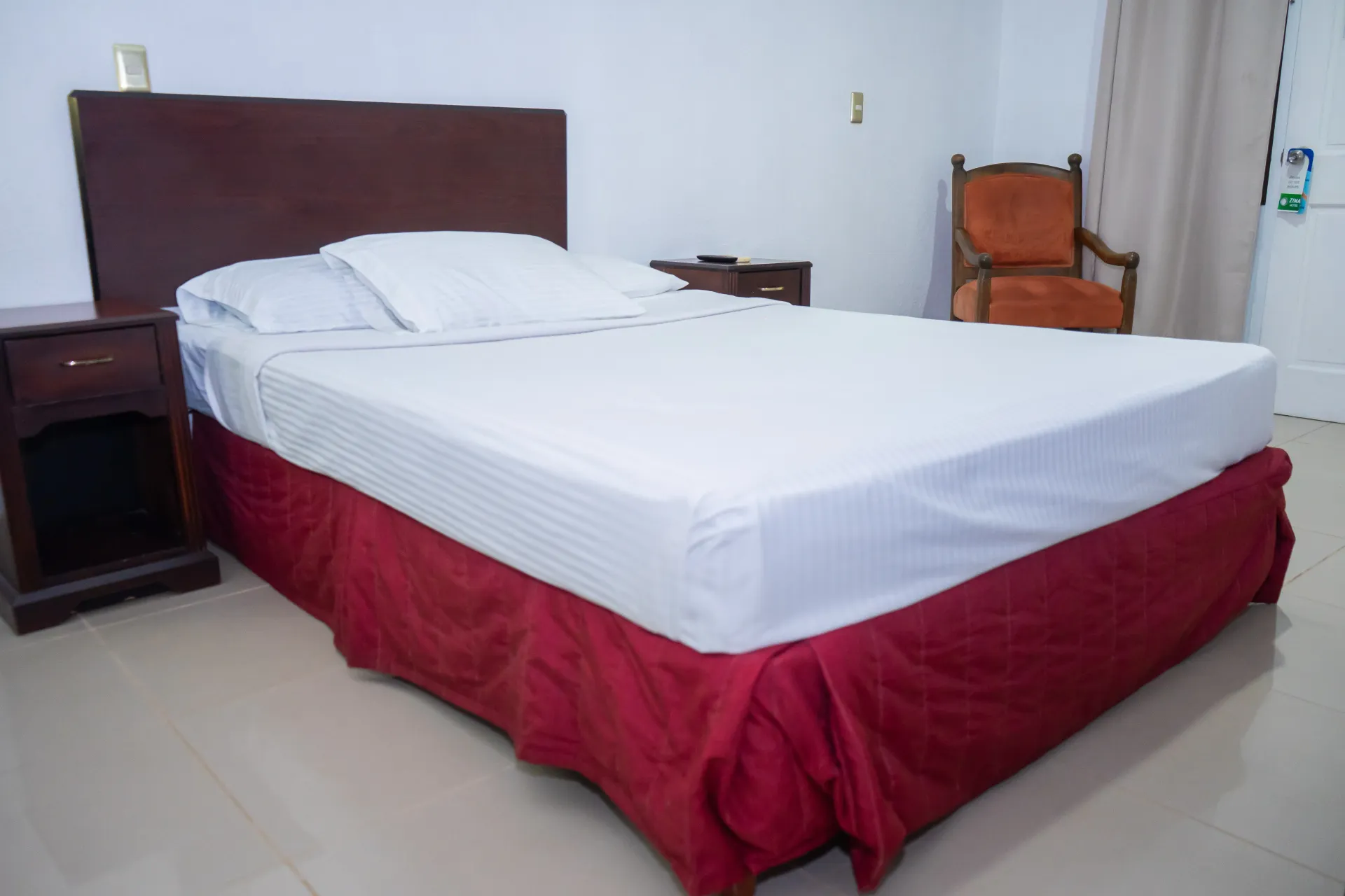 Dormitorio con cama, mesitas de noche, silla y cortina; la cama tiene sábanas blancas y faldón rojo.