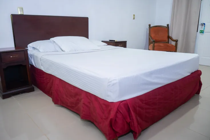 Dormitorio con cama, mesitas de noche, silla y cortina; la cama tiene sábanas blancas y faldón rojo.