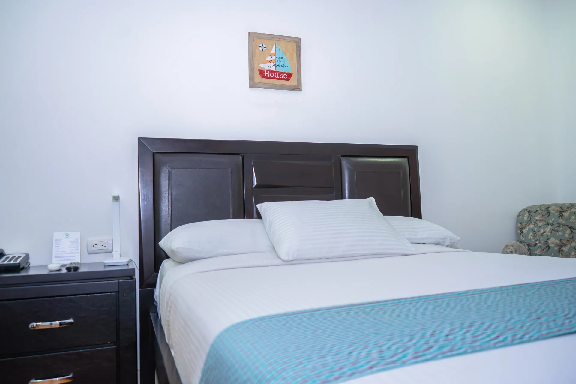 Dormitorio con cama, mesita de noche y una pequeña obra de arte. La cama tiene una alfombra azul y ropa de cama blanca.