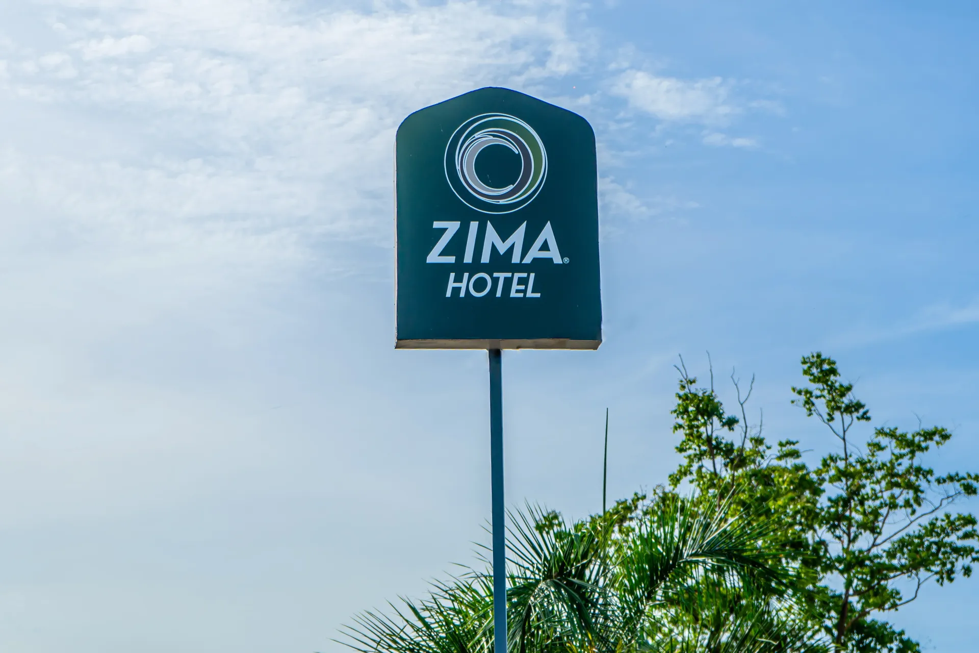 Cartel del Hotel Zima, de color verde oscuro con texto y logotipo en blanco, sobre un cielo nublado.