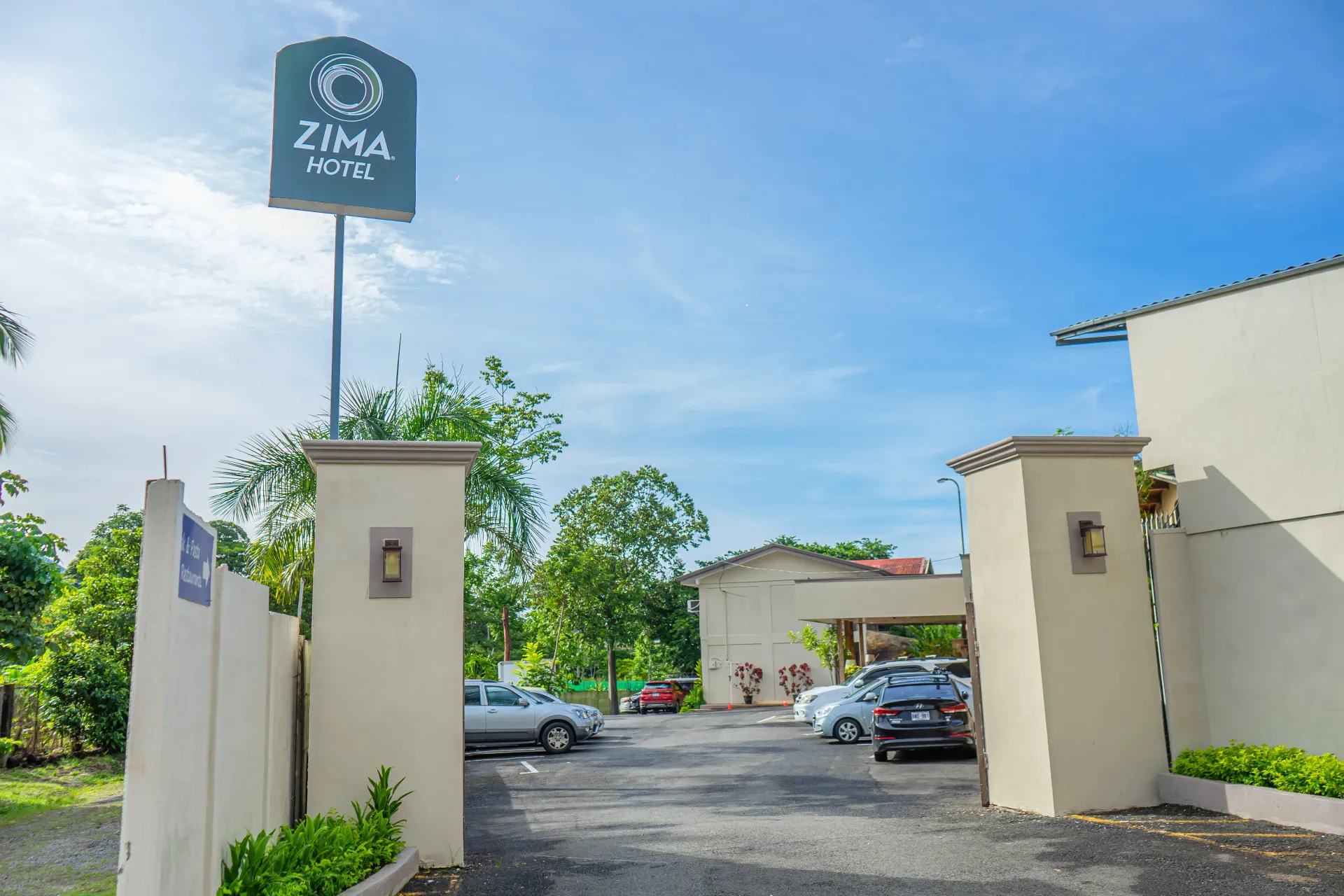 Entrada a Zima Lodge con cartel, pilares, autos estacionados y cielo azul.