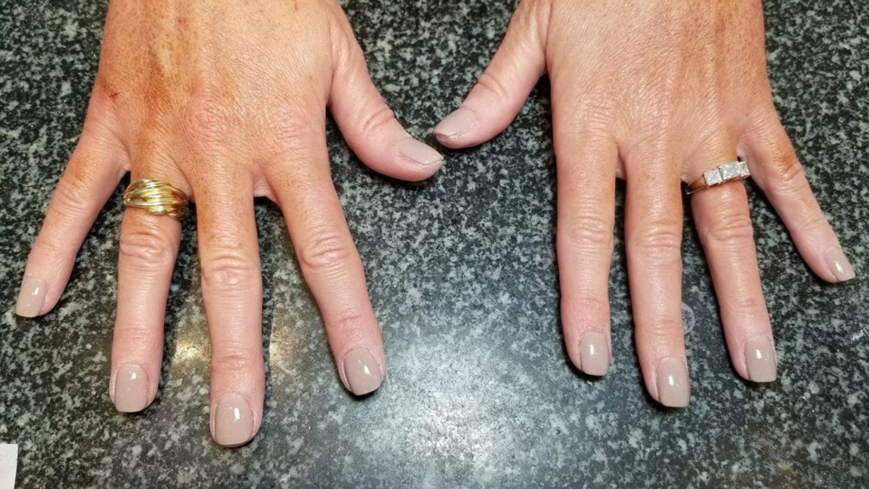 Two hands with painted taupe nails, each wearing rings. Skin shows signs of age.