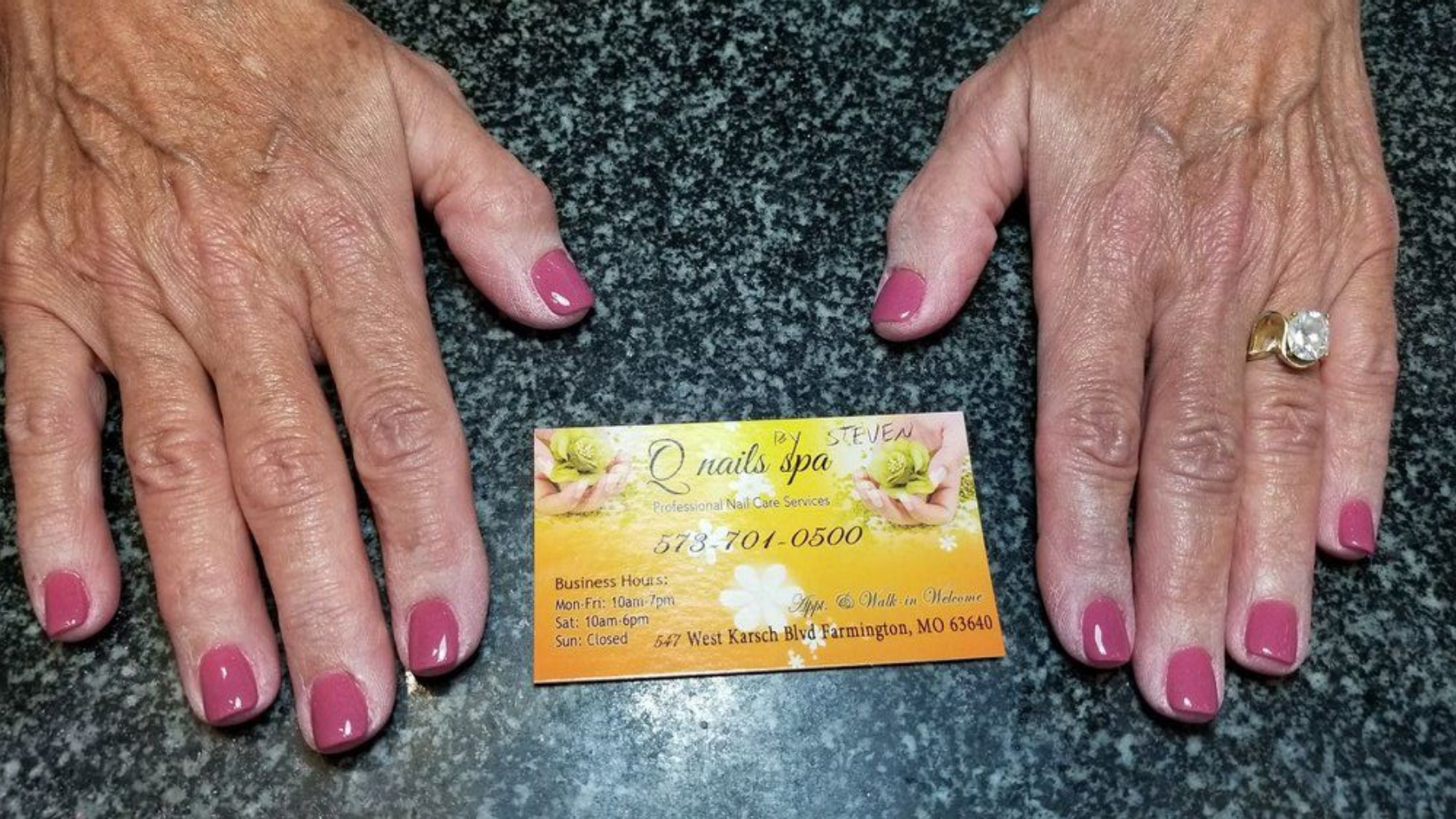 Hands with painted pink nails and business card on a dark surface.