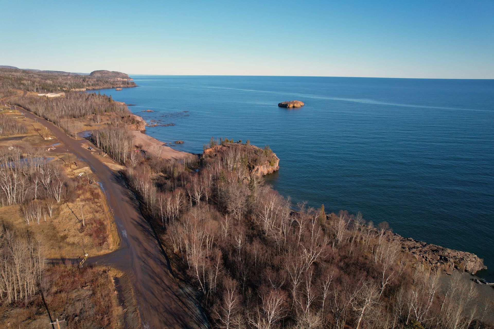 Coastal view with rocky shore, road, bare trees, and blue water under a clear sky.