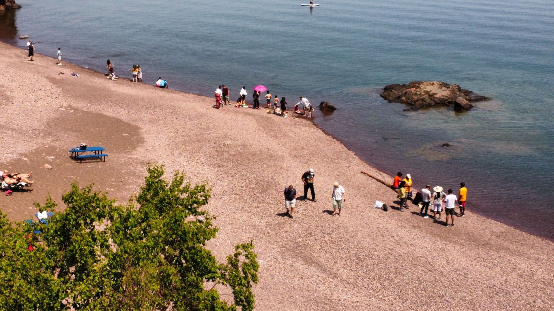 People gather on a rocky beach next to calm blue water on a sunny day.