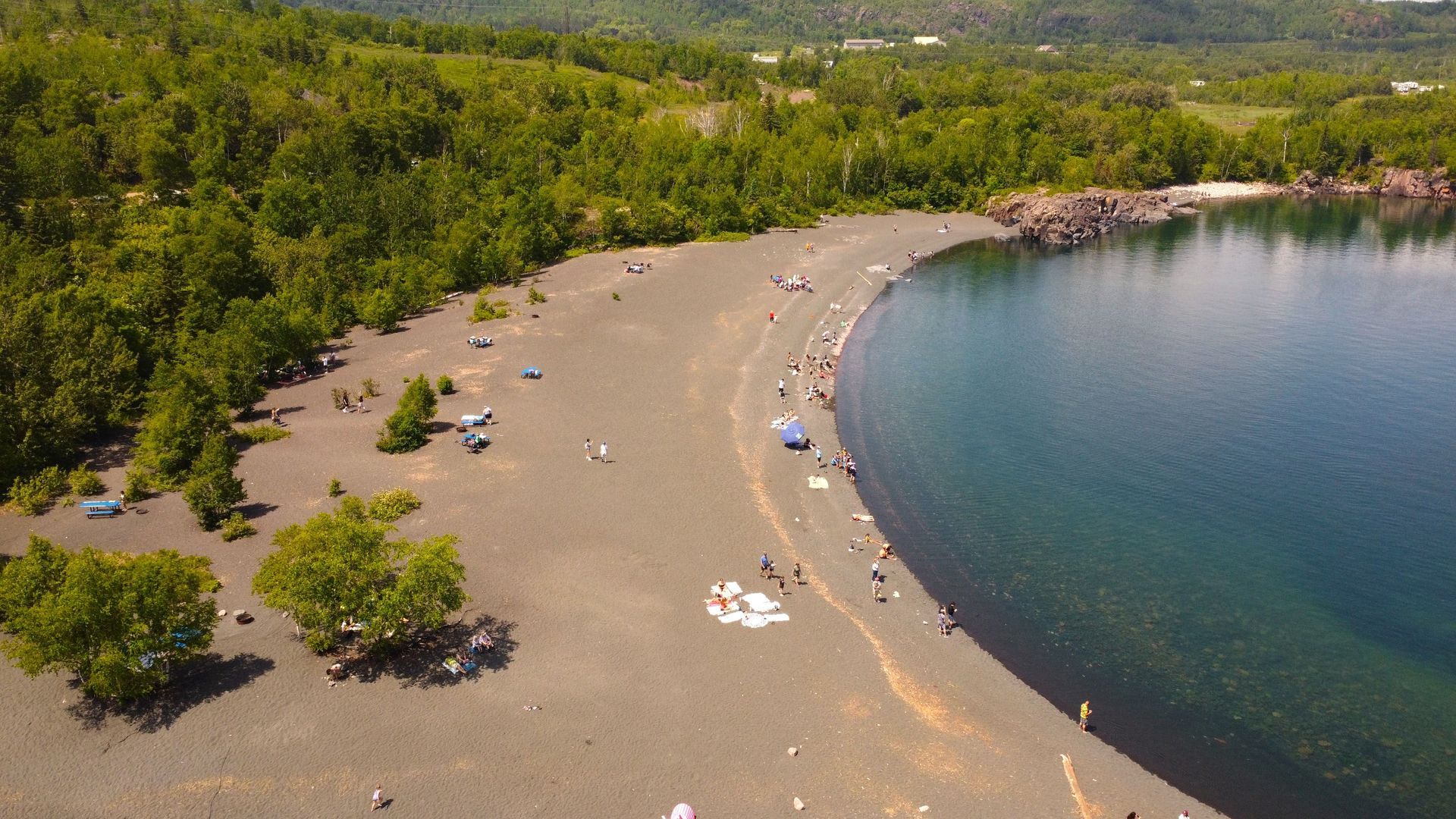 Black sand beach with people, surrounded by dark green forest and clear blue water.