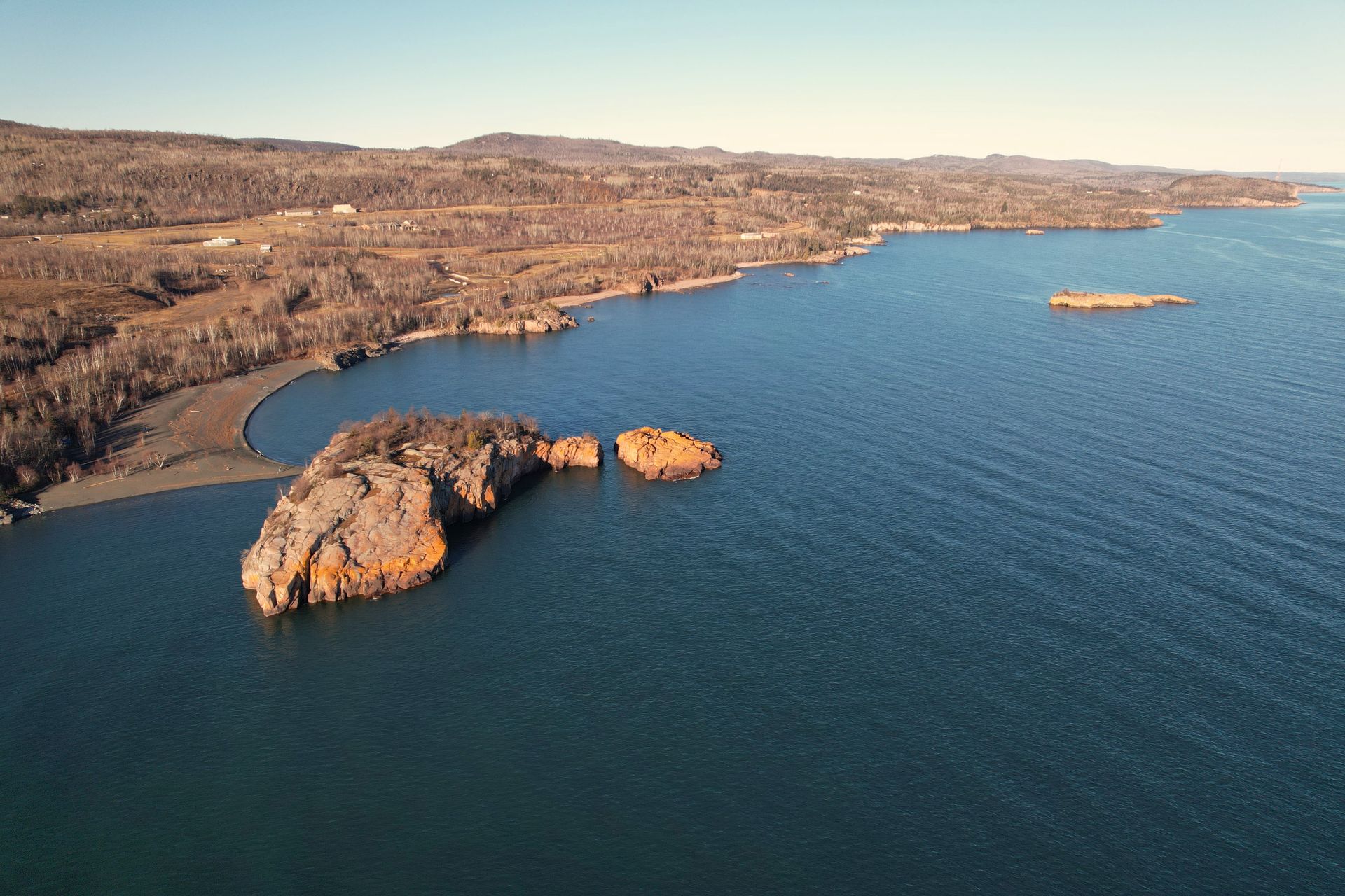 Rocky shoreline and island on a calm, blue lake with land in the background under a clear sky.