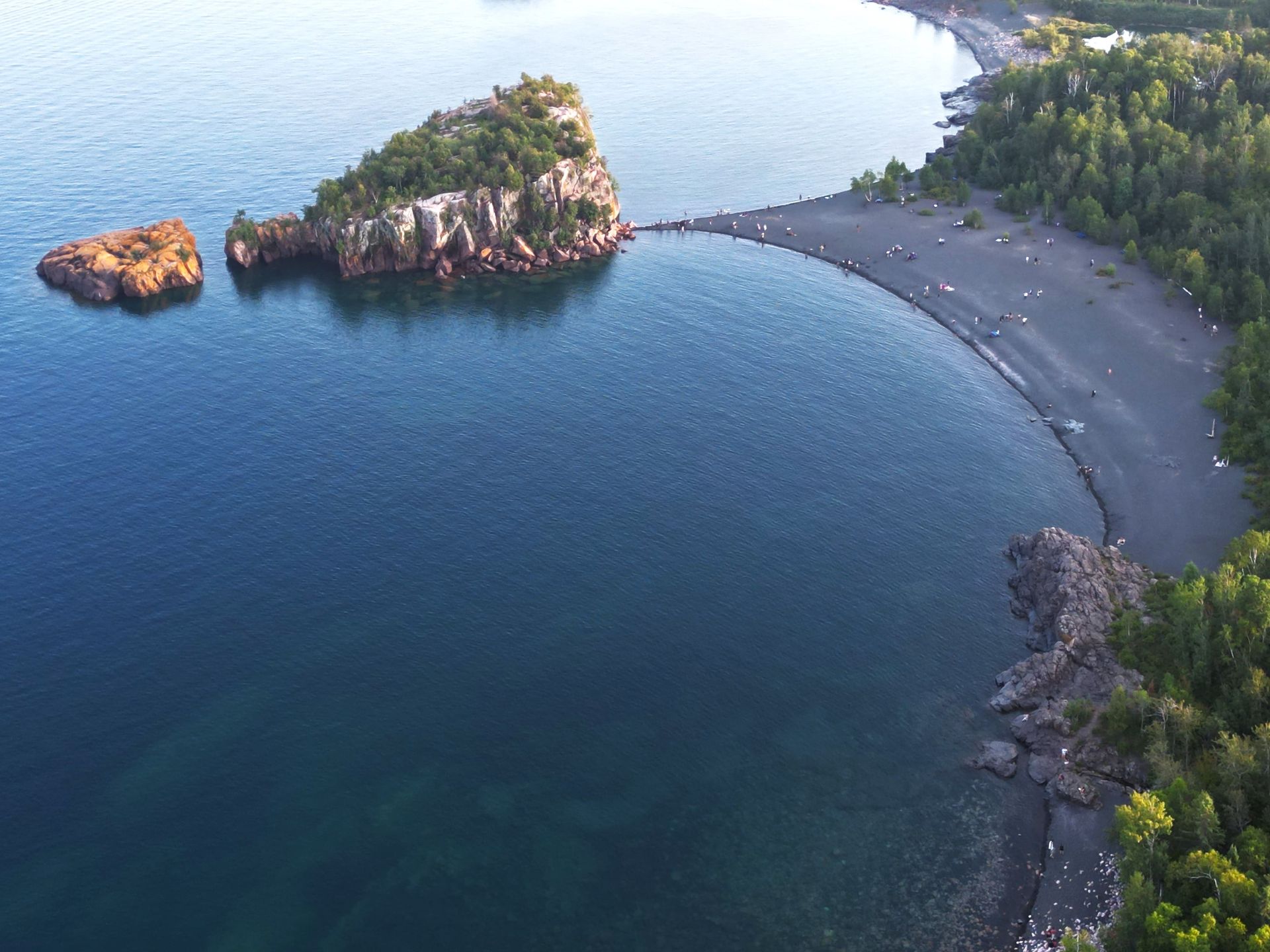 Aerial view of a dark blue bay with a black sand beach and rock formations. Forest surrounds the coastline.