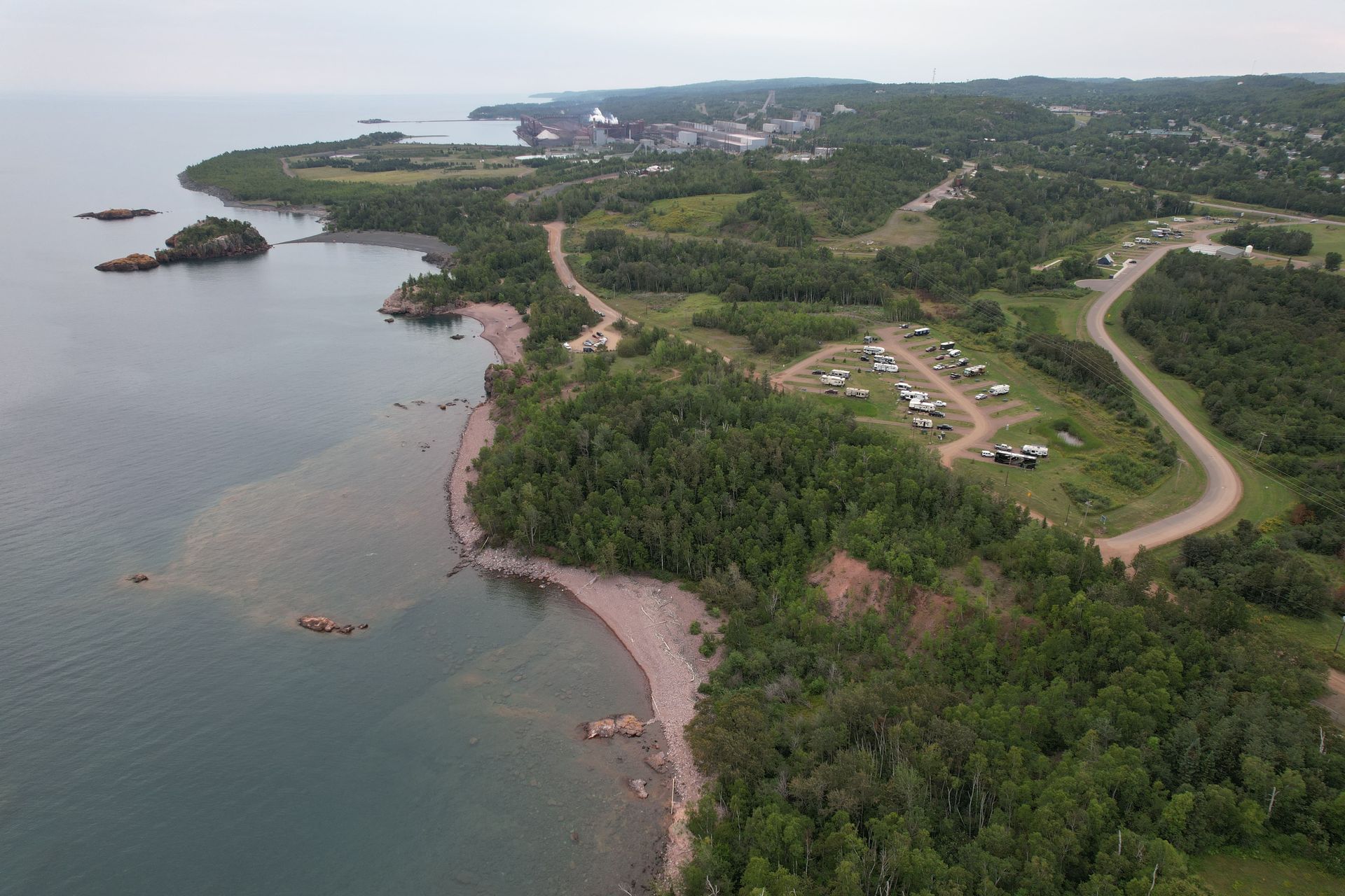 Coastal landscape with rocky shore, green trees, campsite, and town in distance.